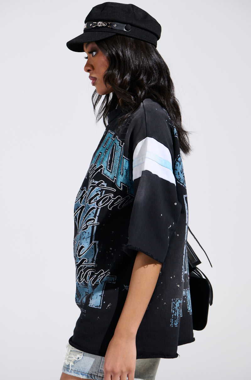 Wearing a black cap and light denim shorts, a person is seen in profile exuding a grungy-chic vibe. They have long dark hair and are carrying a small black bag while dressed casually in the BRIGHT FUTURE OVERSIZED GRAPHIC TEE with blue and white graphics against a plain background.