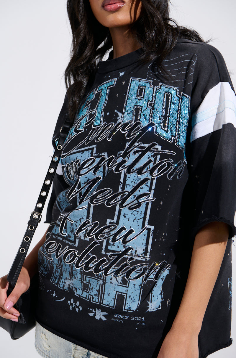 A person with long dark hair wears the BRIGHT FUTURE OVERSIZED GRAPHIC TEE, a grungy-chic black shirt featuring blue and white text like "Revolution" with decorative patterns, holding a black strap over their shoulder.