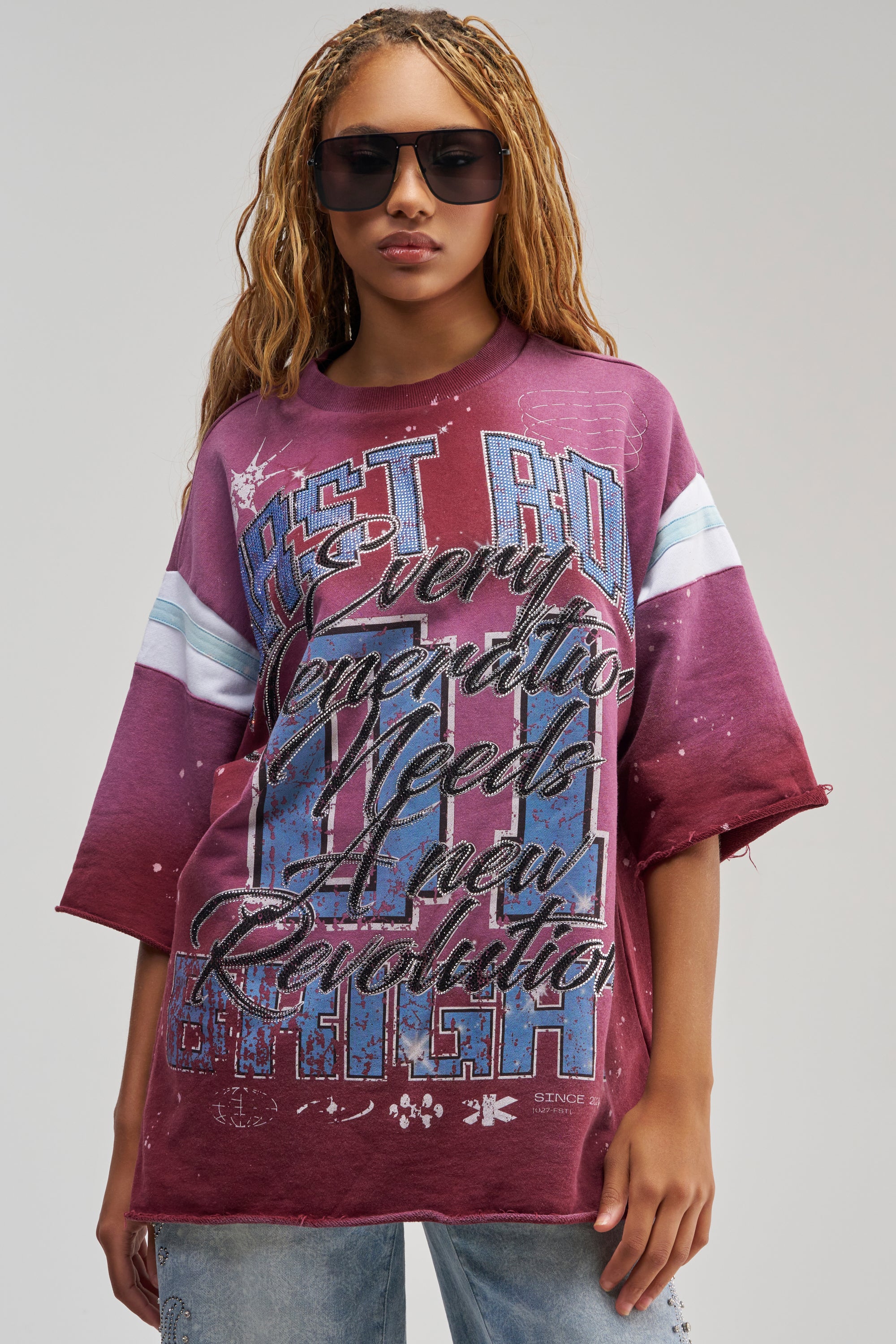 A person wearing sunglasses and the MY FUTURE OVERSIZED GRAPHIC TEE IN BURGUNDY, which features bold blue and white text that reads “Every Generation Needs a New Revolution,” stands facing forward. The loose-fit tee exudes a strong streetwear vibe, complemented by their hair styled in relaxed waves.