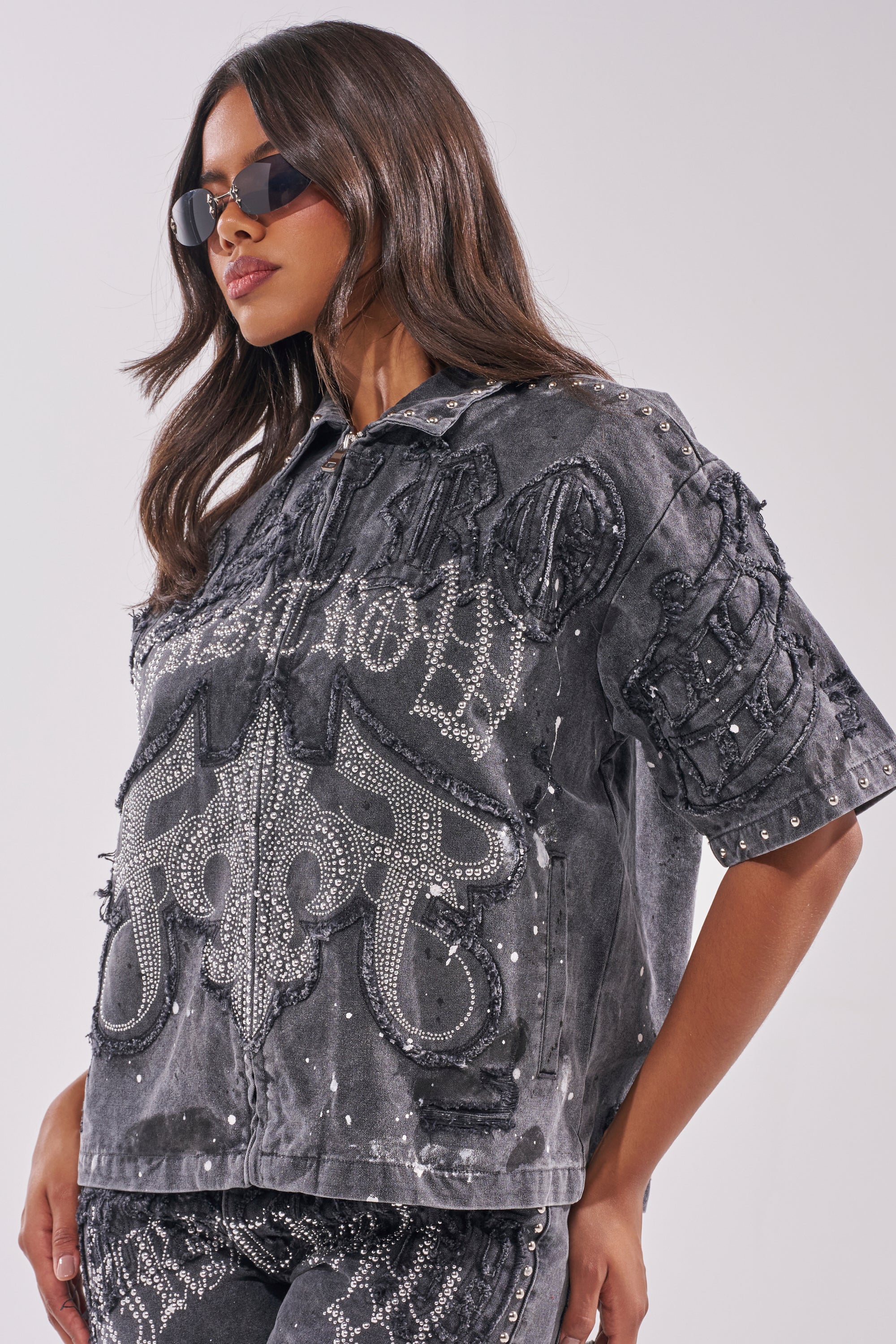 A woman wearing black sunglasses and the NOT LIKE THE REST EMBELLISHED TOP with silver studs and intricate patterns poses against a plain light background.
