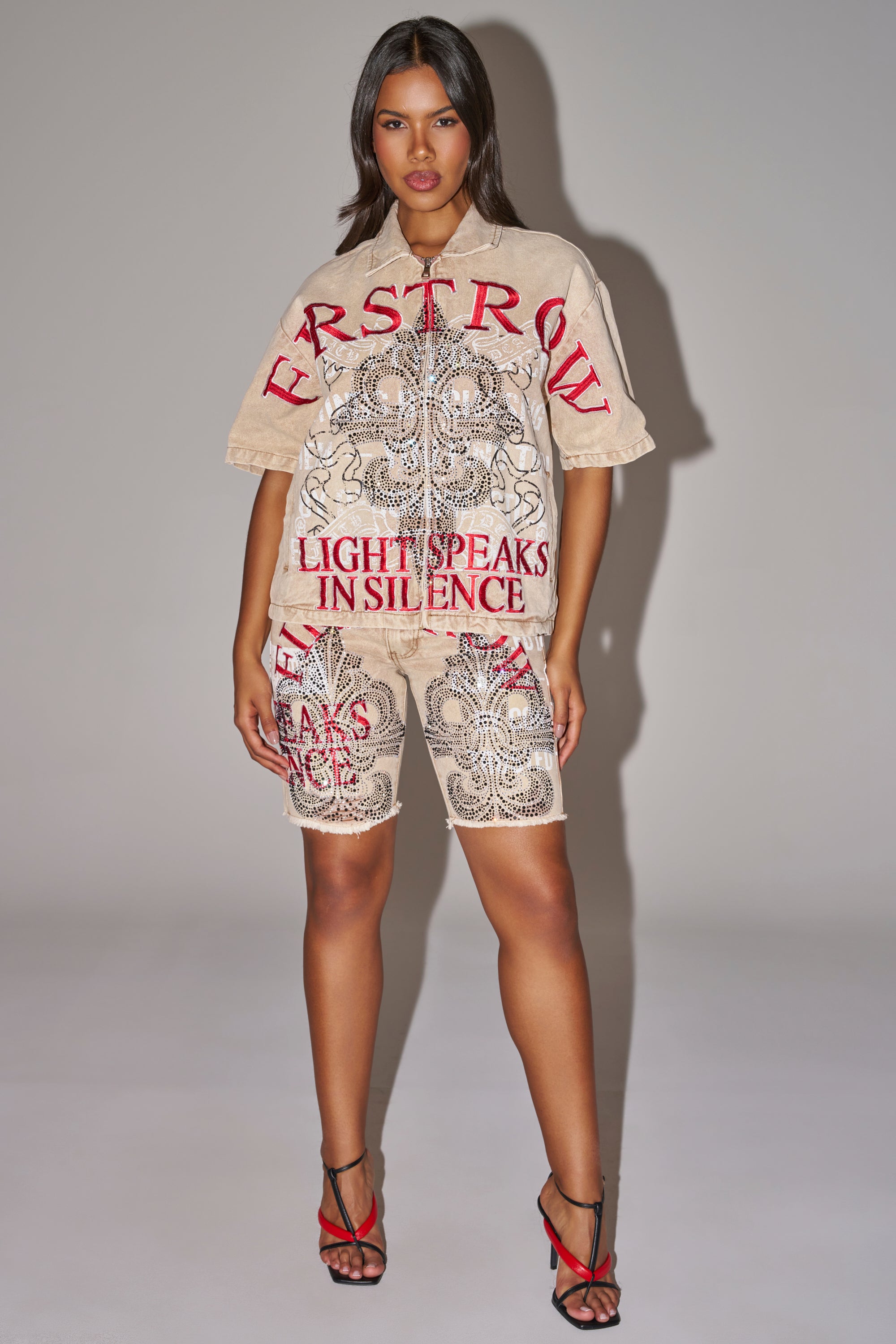A woman stands confidently in the LIGHT SPEAKS IN SILENCE CANVAS SHIRT and matching shorts with embroidered rhinestones, red text, and black designs stating “FIRSTROW” and “LIGHT SPEAKS IN SILENCE.” She pairs the look with strappy heels. 9