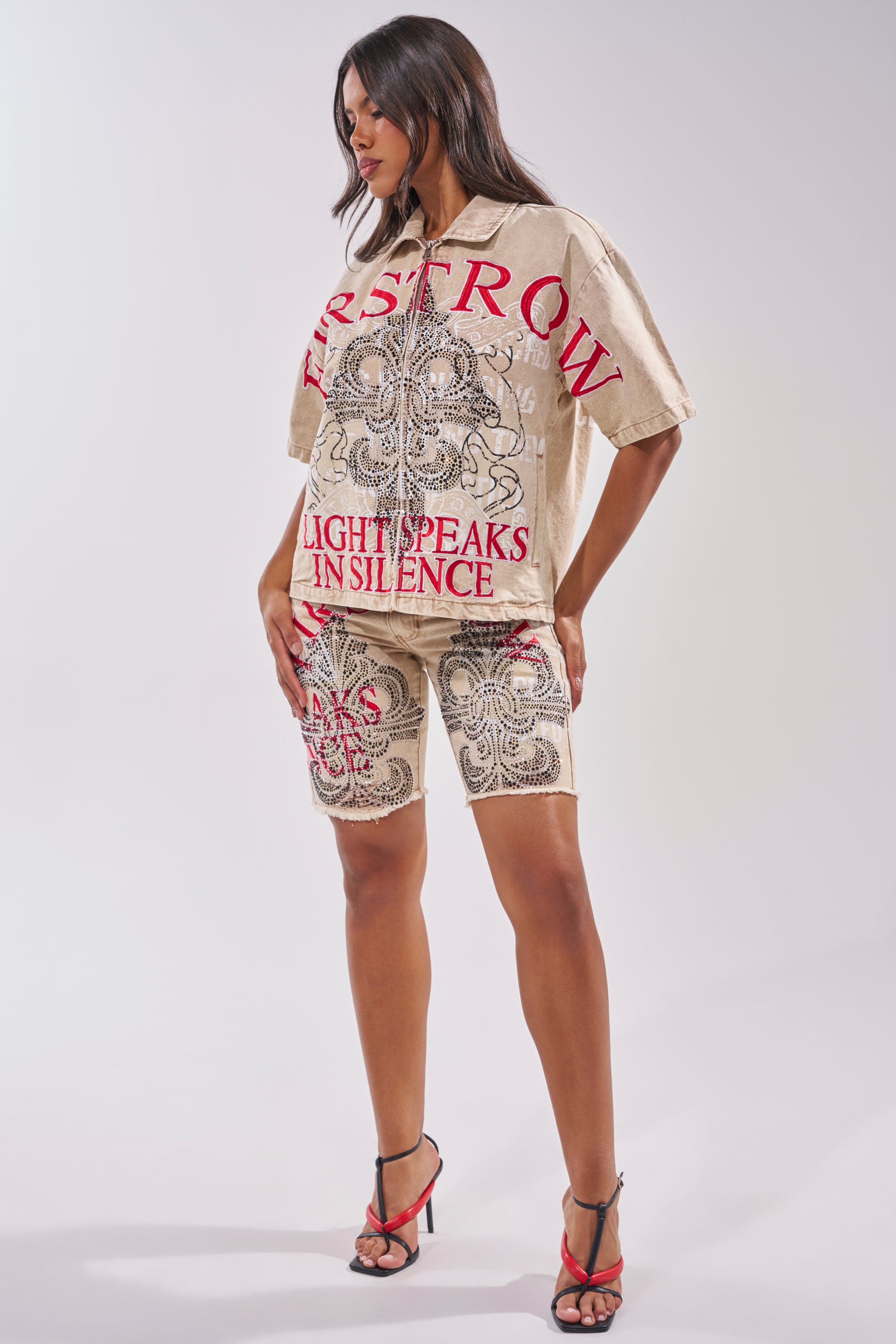 A woman with long hair wears the LIGHT SPEAKS IN SILENCE CANVAS SHIRT—a light brown denim shirt featuring bold red text and detailed black designs—paired with matching shorts and black and red high-heeled sandals. 11