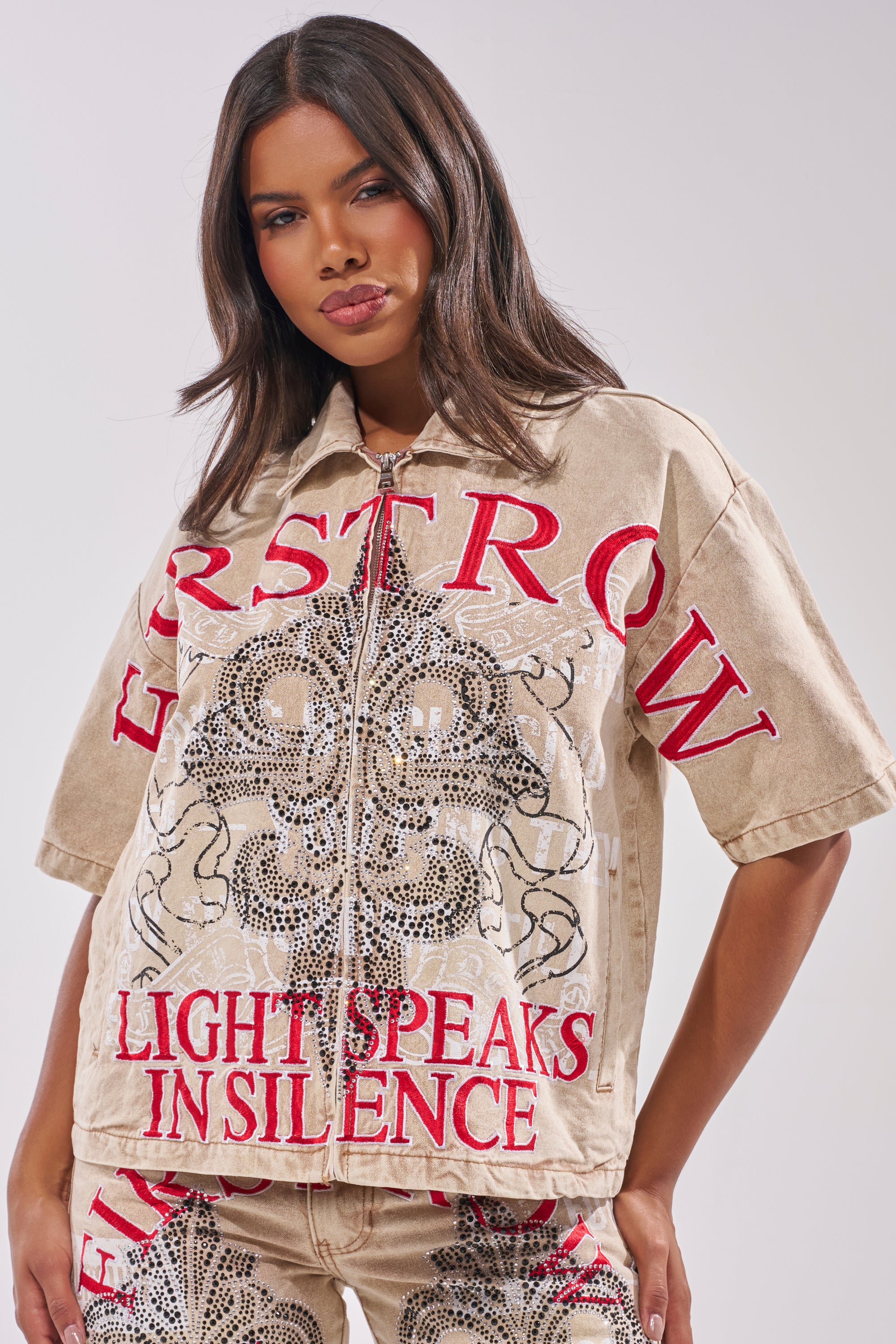 A woman with long dark hair wears the LIGHT SPEAKS IN SILENCE CANVAS SHIRT and matching pants, both light-brown with bold red "FIRSTROW" and "LIGHT SPEAKS IN SILENCE" text, plus intricate studded and graphic details. 1