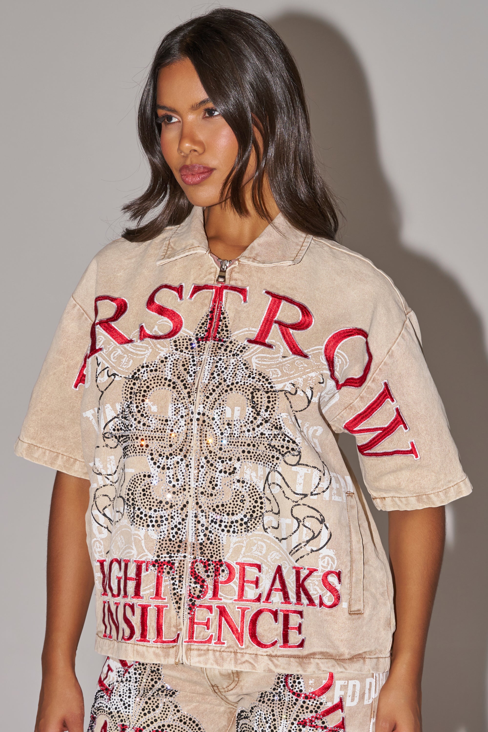 A woman wears the LIGHT SPEAKS IN SILENCE CANVAS SHIRT—a light-brown denim shirt with red embroidered lettering, skull designs, and rhinestone accents—standing against a plain background. 2