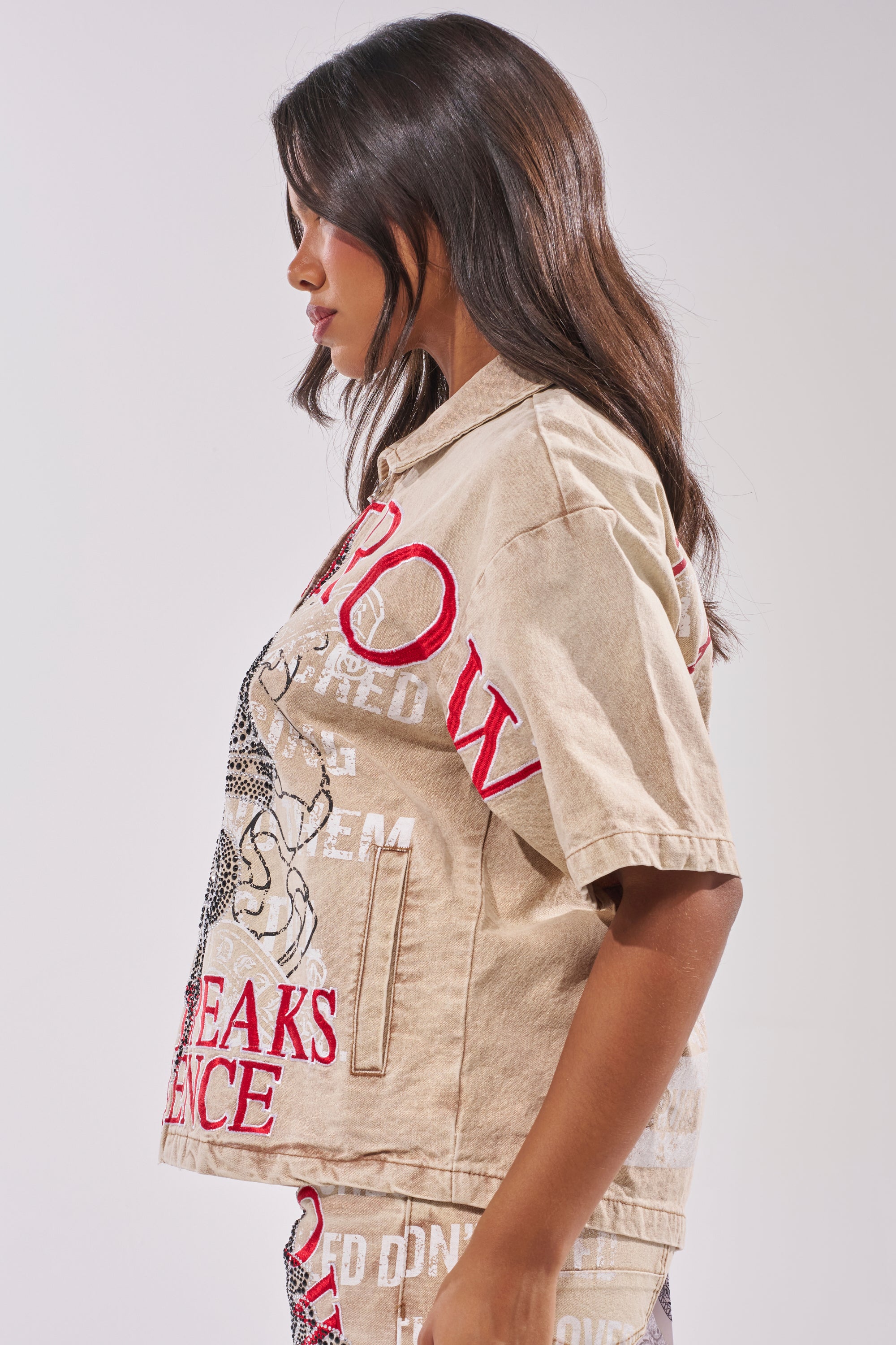 A woman with long, dark hair stands in profile wearing the LIGHT SPEAKS IN SILENCE CANVAS SHIRT, an oversized relaxed-fit top featuring red and white text and graphics, against a plain light-colored background. 5