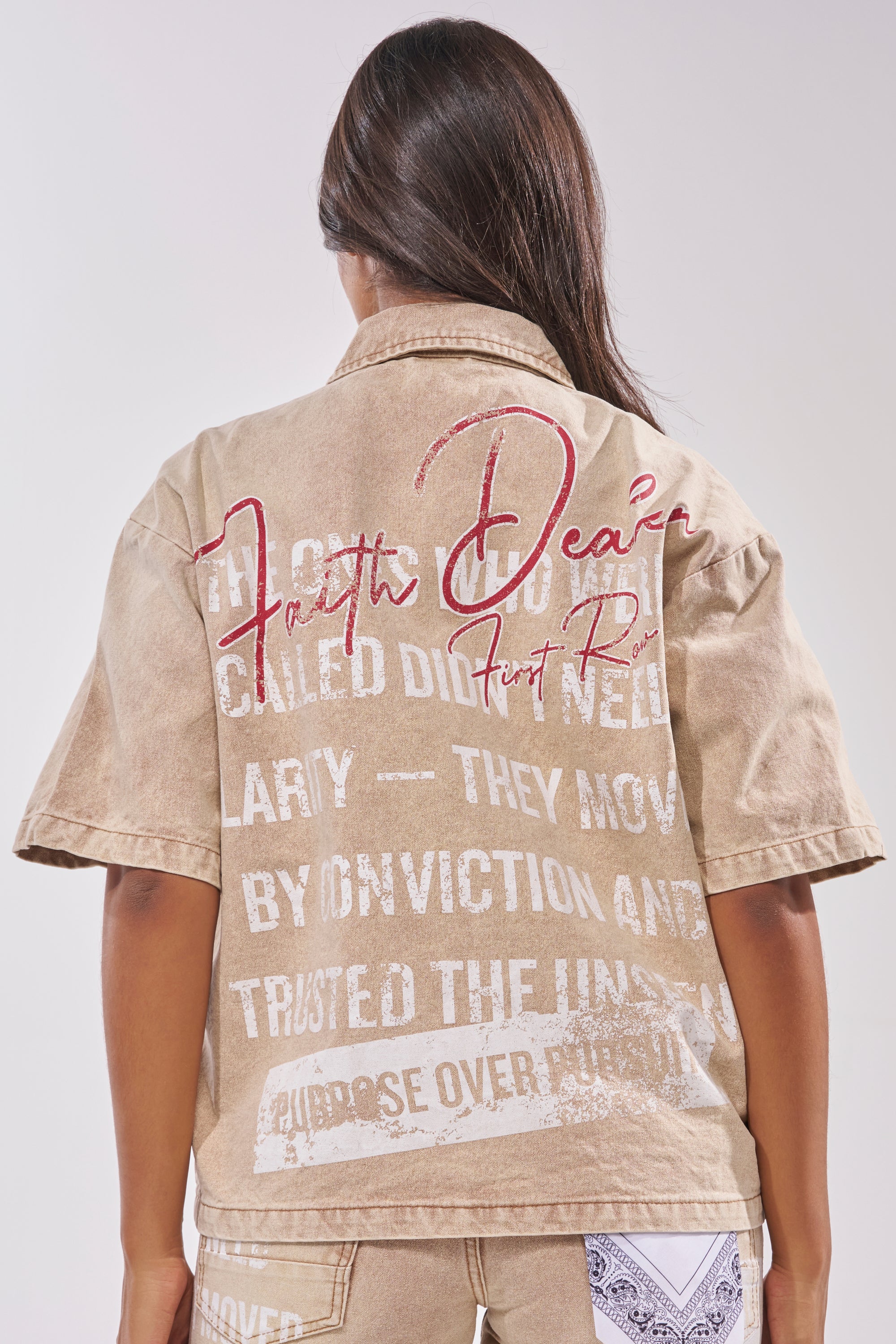 A person with long brown hair is seen from the back, wearing the LIGHT SPEAKS IN SILENCE CANVAS SHIRT—a light brown denim shirt featuring white and bold red script along with block letters in various sizes and styles. 6
