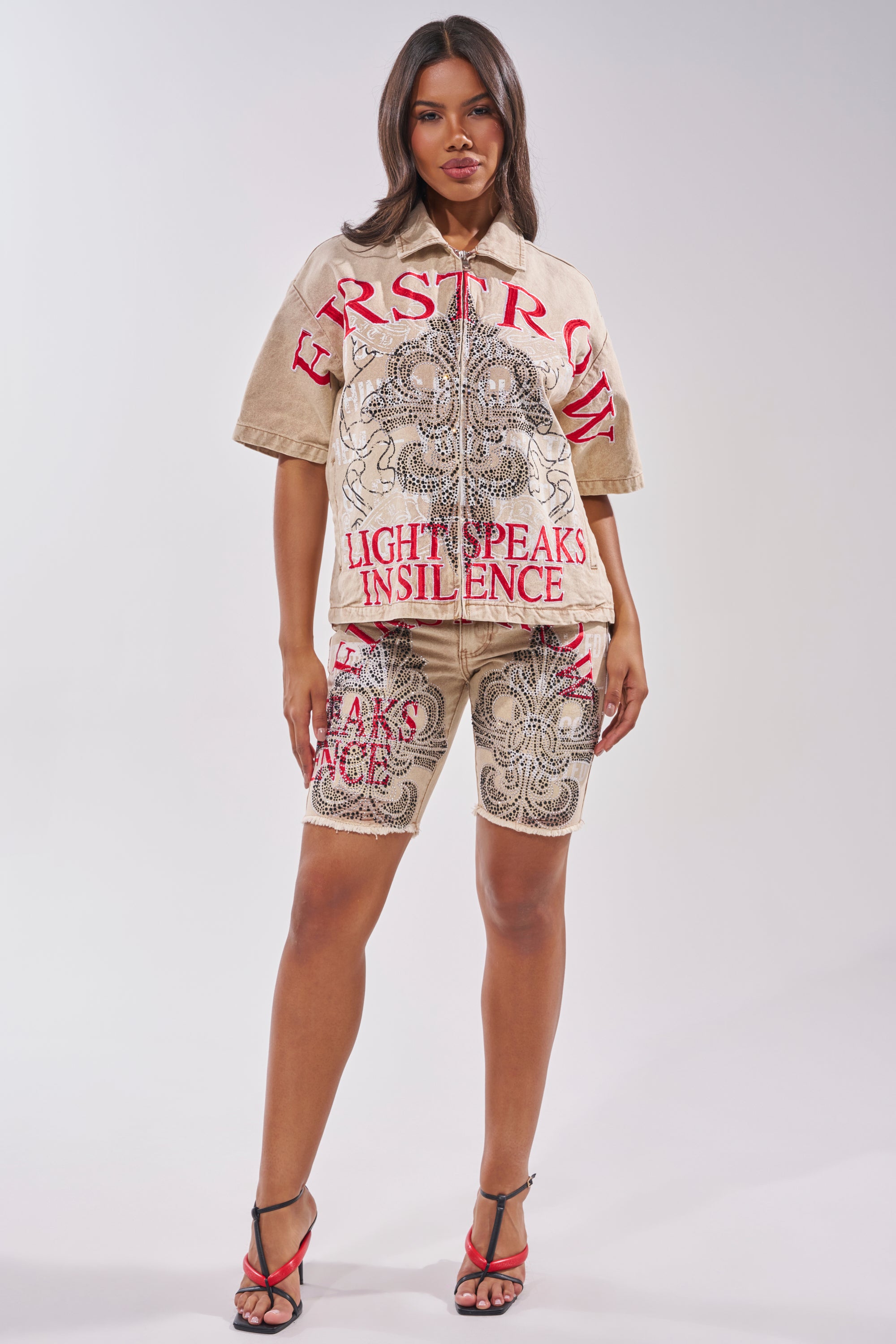 A woman wears the LIGHT SPEAKS IN SILENCE CANVAS SHIRT, a tan top with lace-like patterns, subtle rhinestones, and bold red text: "FIRST ROW" and "LIGHT SPEAKS IN SILENCE." She pairs it with matching shorts and strappy black and red heels. 8