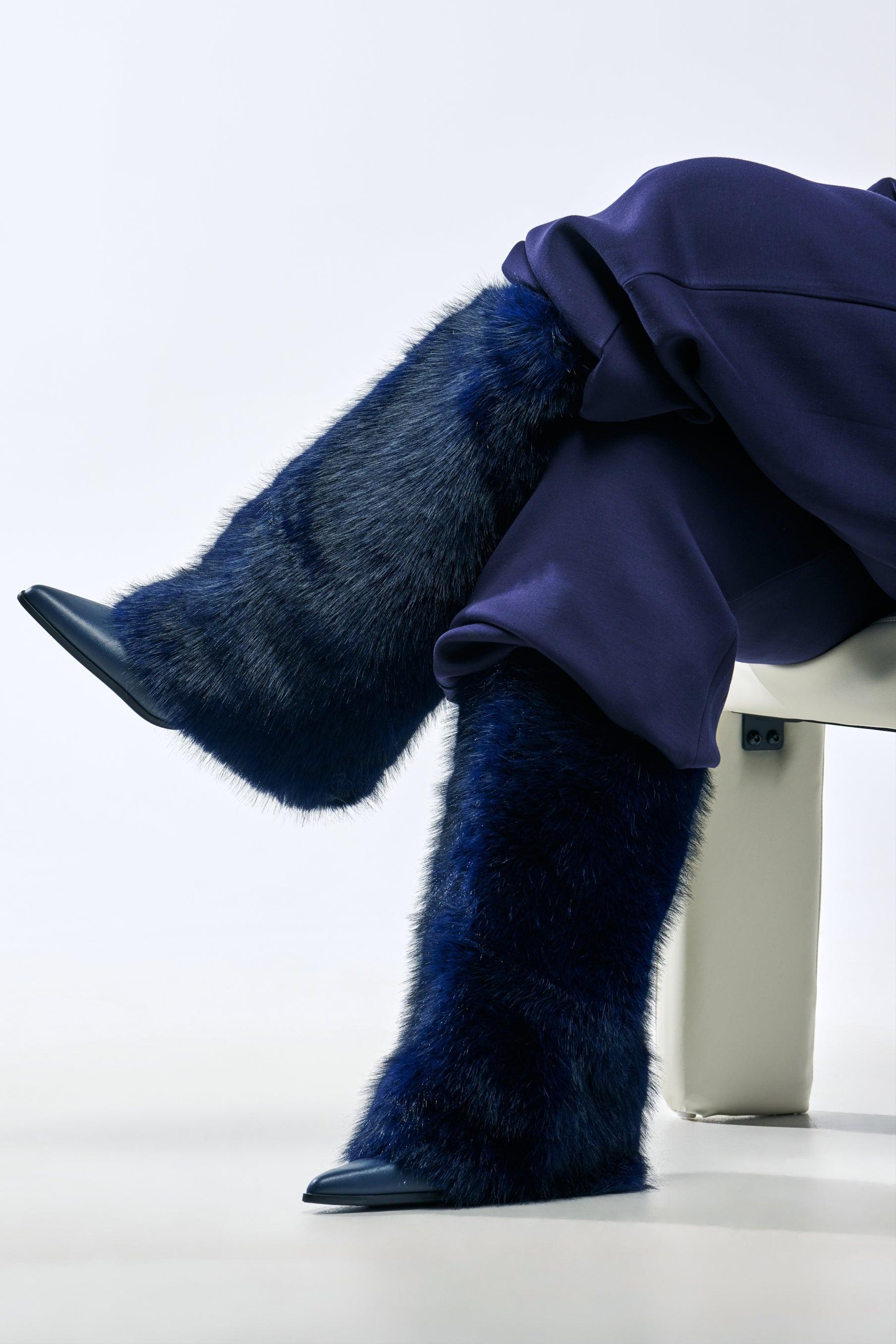 A person sits with legs crossed wearing AZALEA WANG FRIATA NAVY FUR WESTERN BLOCK BOOT knee-high navy faux fur cowboy boots with pointed toes.