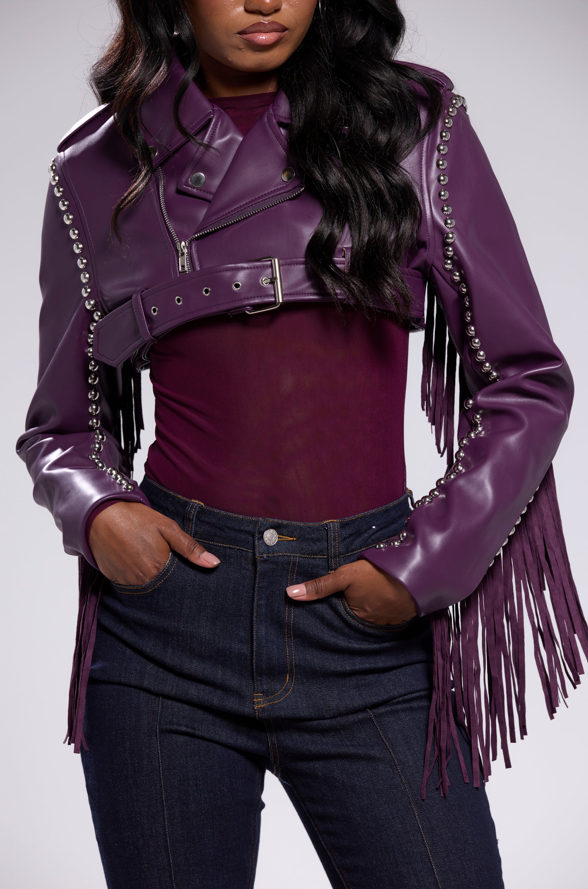 A woman wears the HOLD EM CROPPED FRINGE MOTO jacket with silver studs over a sheer purple top and high-waisted dark jeans, posing with one hand in her pocket. 10