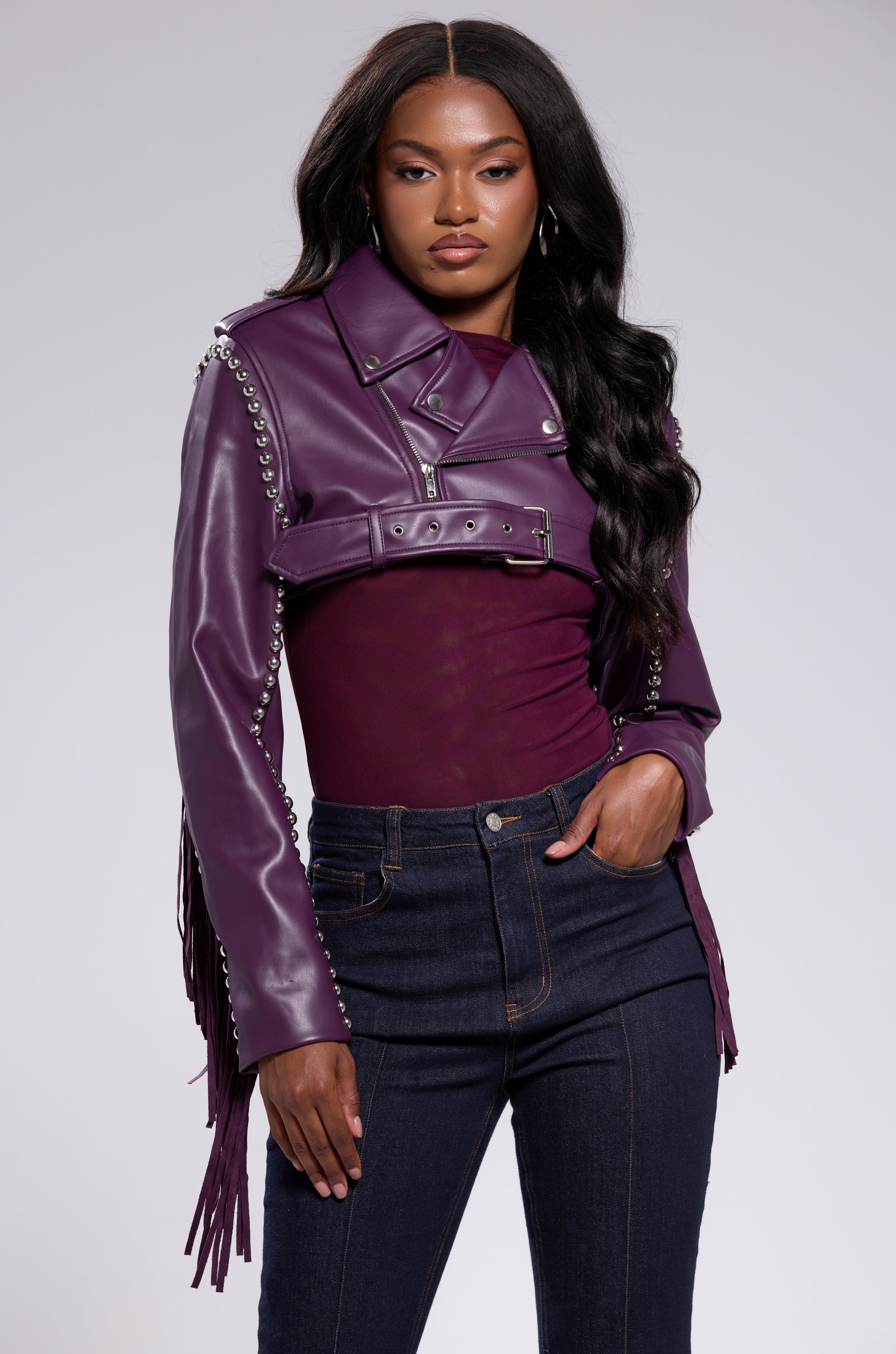 A woman stands confidently against a plain light gray background wearing the HOLD EM CROPPED FRINGE MOTO by AKIRA Label, featuring silver studs, over a maroon top and dark jeans. 1