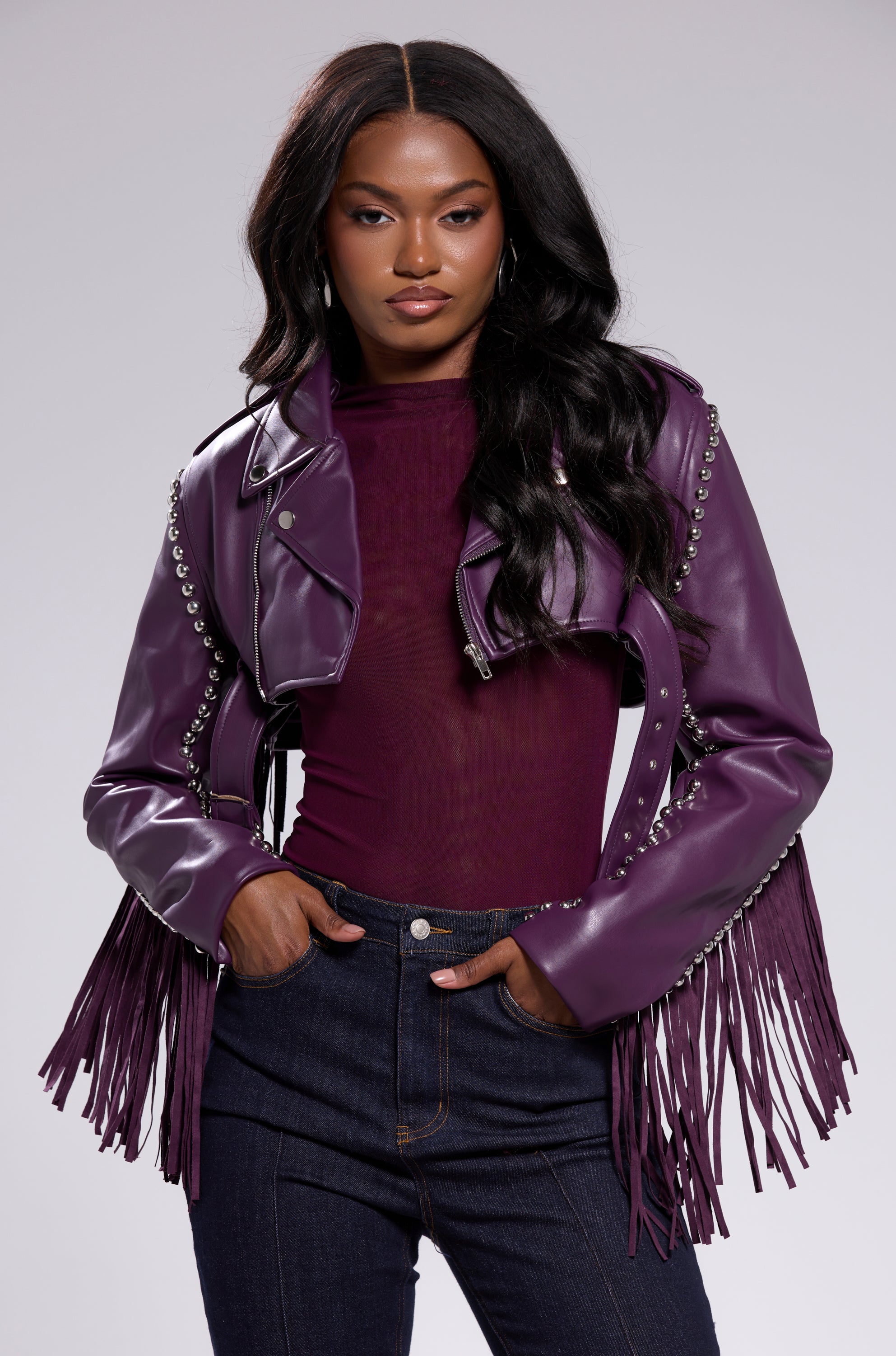 A woman with long black hair poses confidently in the HOLD EM CROPPED FRINGE MOTO jacket, styled over a matching top and dark jeans, one hand in her pocket against a plain background. 2