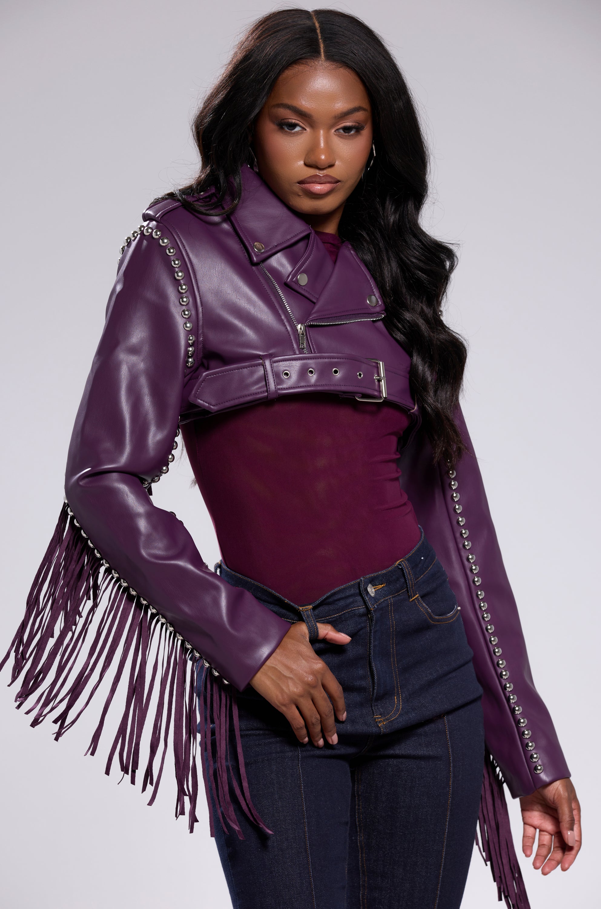 A woman poses confidently in the HOLD EM CROPPED FRINGE MOTO jacket, styled with a matching purple top and dark high-waisted jeans, set against a light gray background. 4