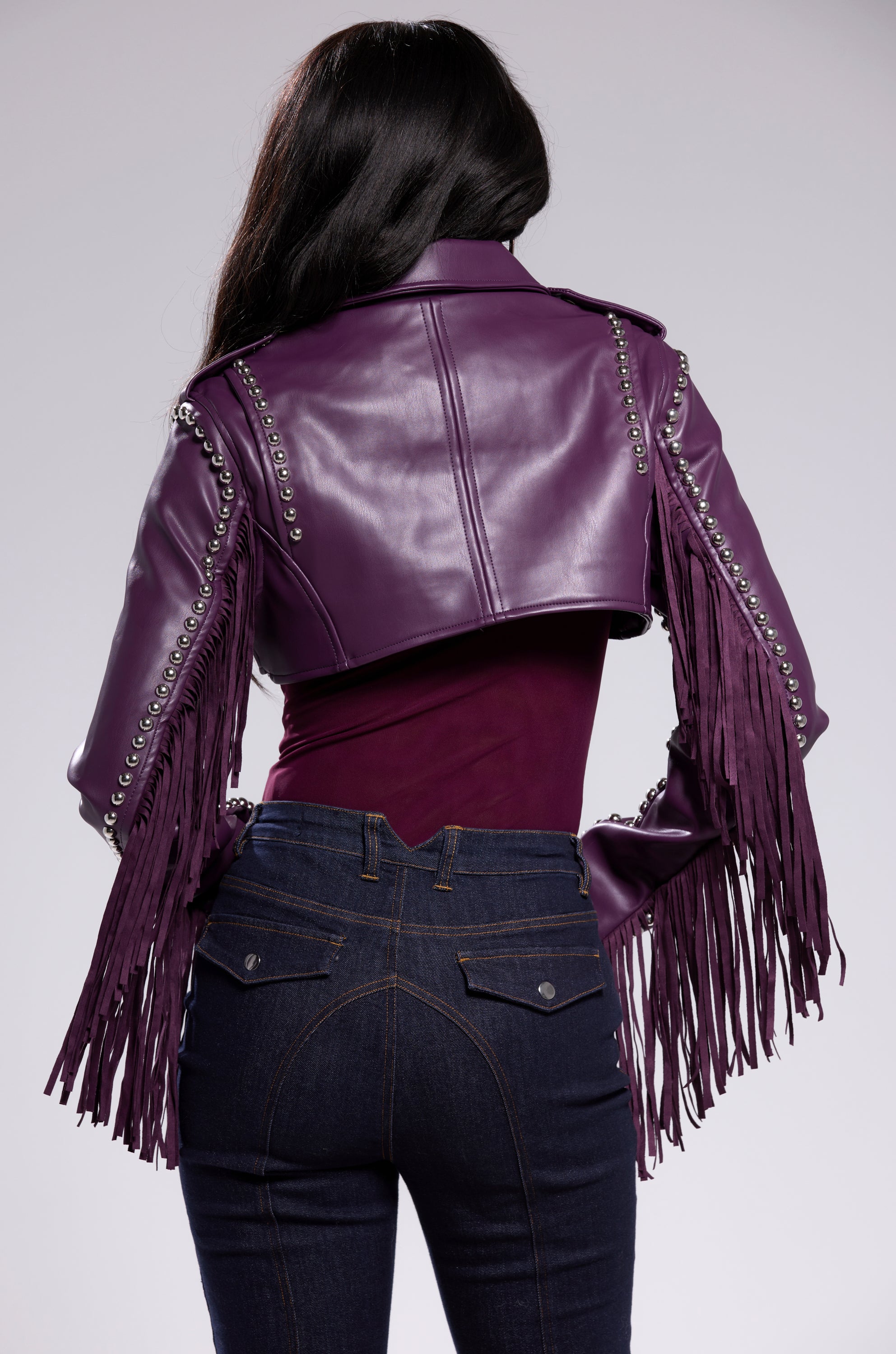 A person with long dark hair, seen from behind, wears the HOLD EM CROPPED FRINGE MOTO jacket with silver hardware over a fitted maroon top and dark blue jeans. 6