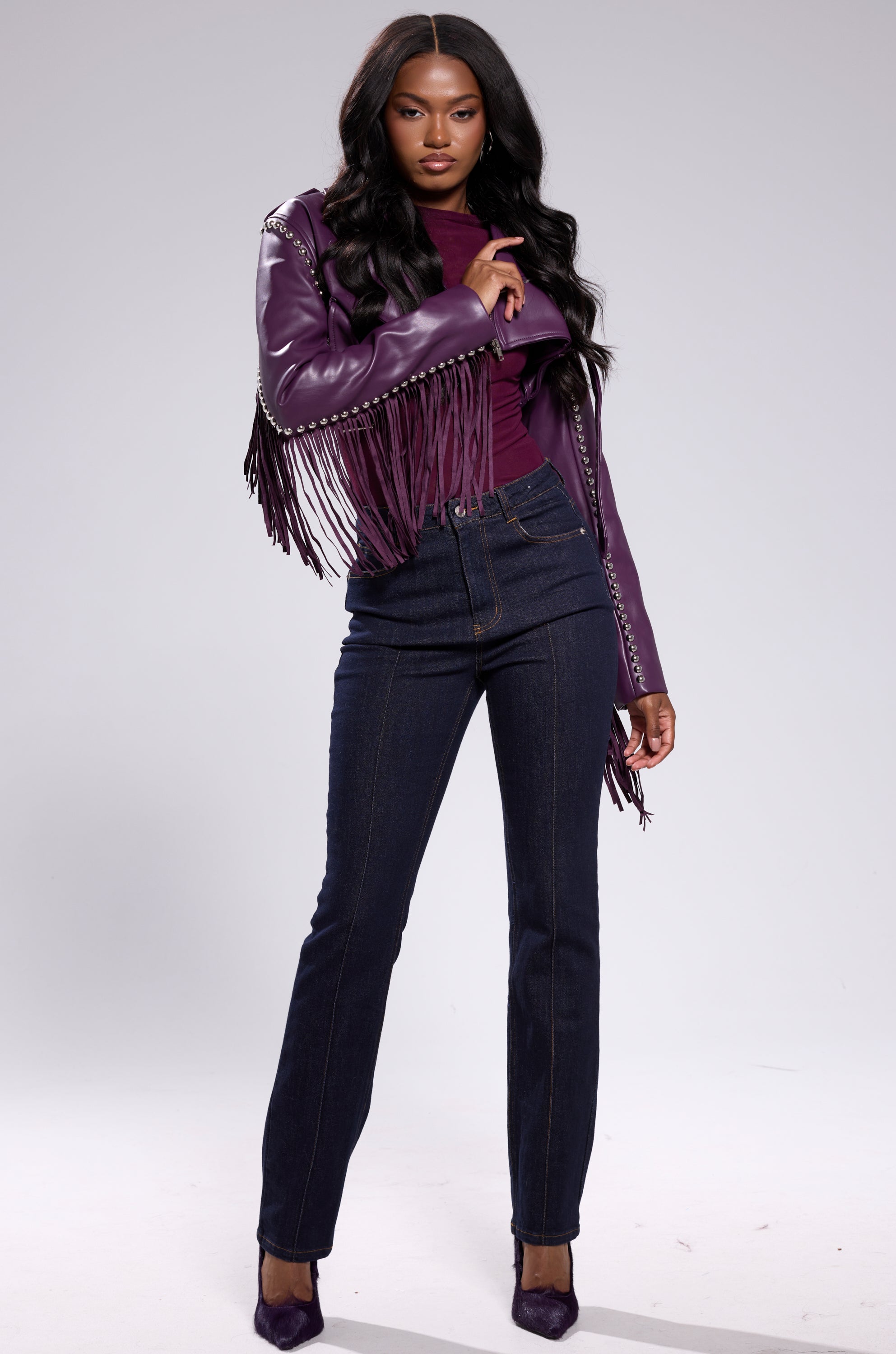 A woman with long dark hair models the HOLD EM CROPPED FRINGE MOTO jacket with silver hardware, a purple top, dark blue high-waisted jeans, and purple heels, standing confidently against a neutral background. 8