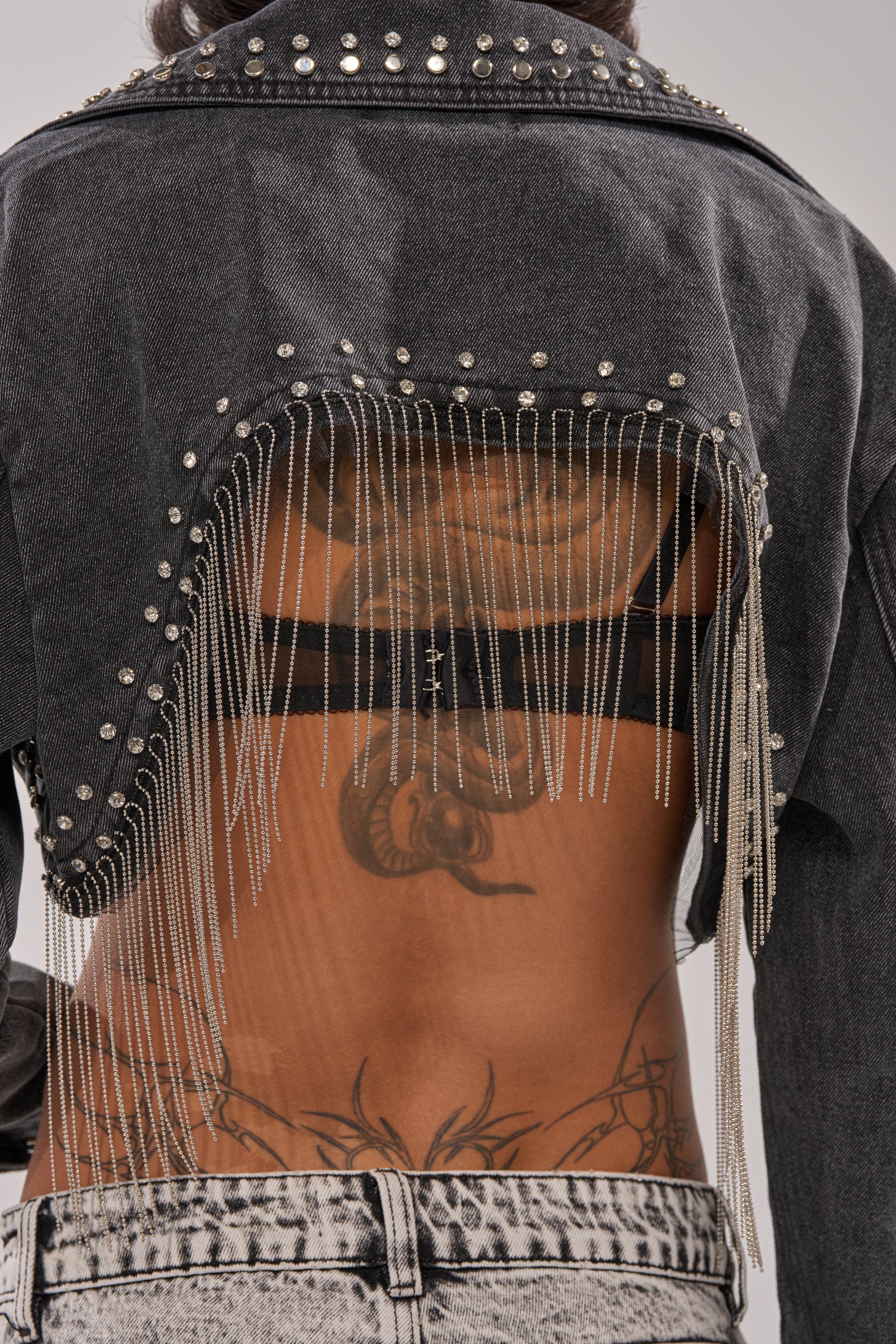Wearing the WATERFALL FRINGE CROPPED DENIM JACKET with beaded fringe and an open back, styled with light-wash jeans to reveal tattoos and a black bra strap for an edgy, bold look. 9