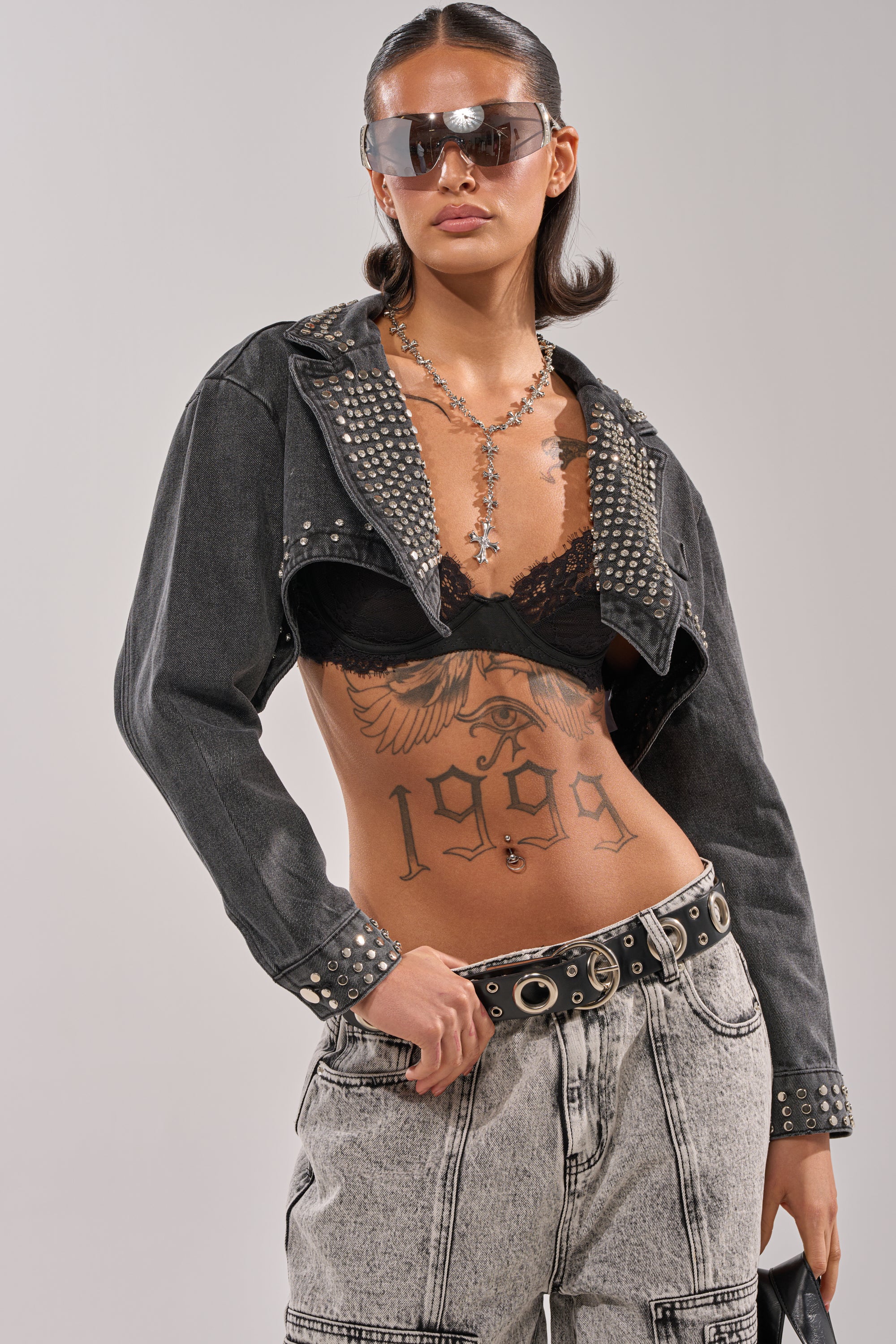 A woman wears the WATERFALL FRINGE CROPPED DENIM JACKET with a black lace bra, light-wash high-waisted jeans, large sunglasses, and a cross necklace. She confidently shows off a bold "1999" tattoo on her torso. 1
