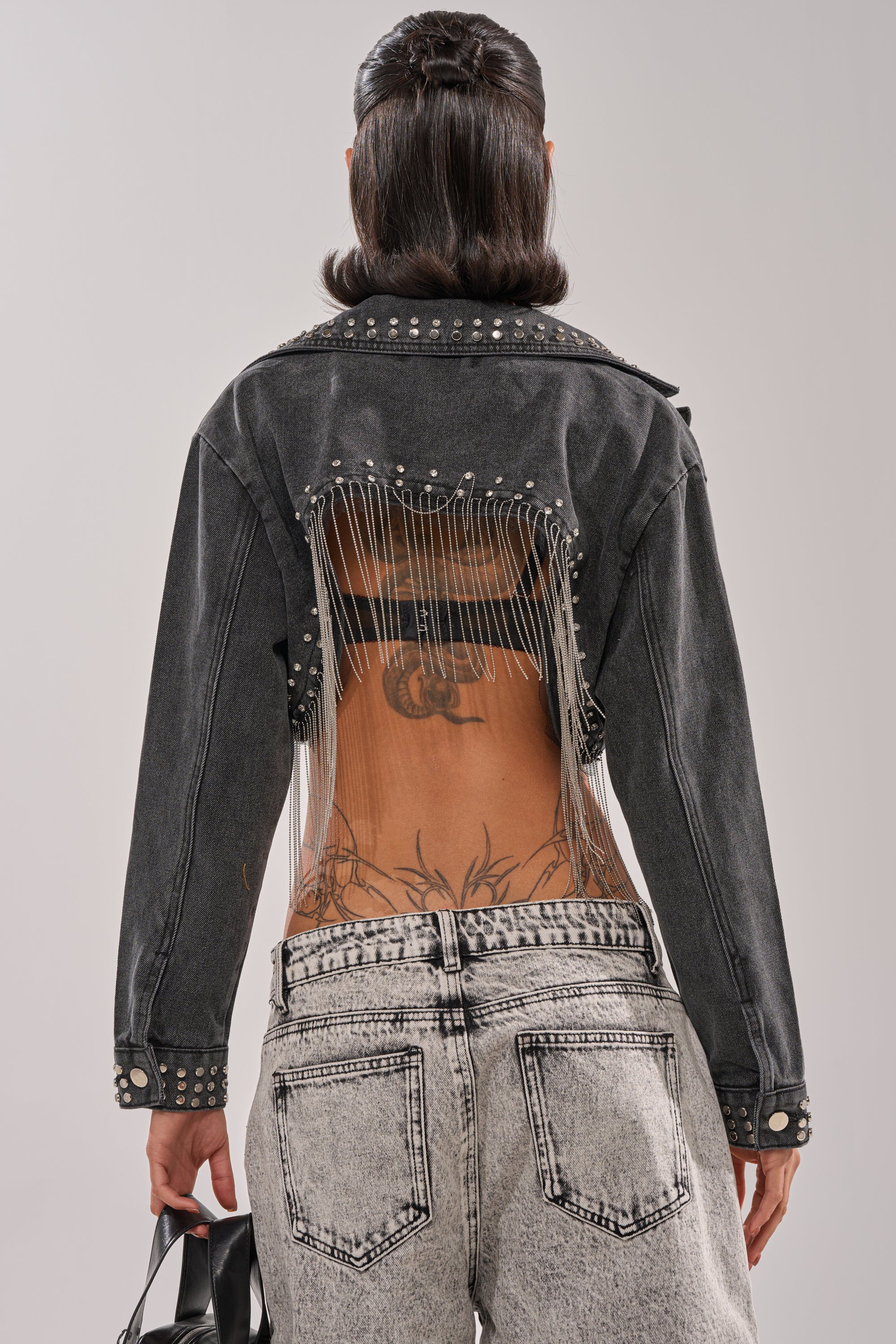 A person with shoulder-length dark hair and lower back tattoos is seen from behind in the WATERFALL FRINGE CROPPED DENIM JACKET, featuring rhinestone studs, vertical chain detail on an open back, and paired with light-washed jeans. 4