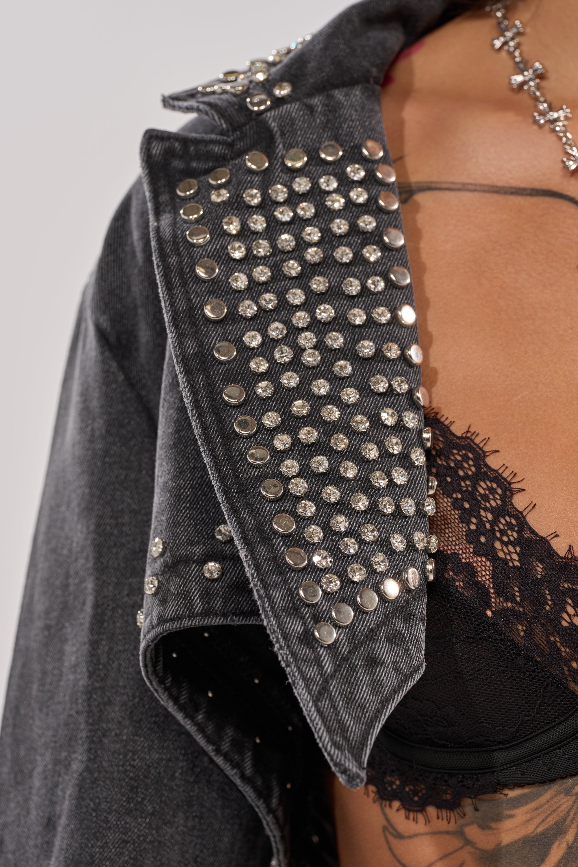 Close-up of a person wearing the WATERFALL FRINGE CROPPED DENIM JACKET with silver-studded collar over a black lace bralette. A partial necklace and chest tattoo are also visible. 8