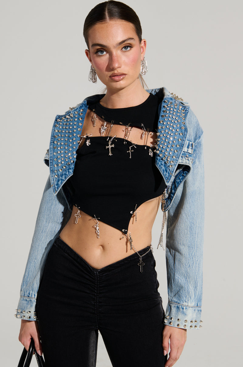 A person wearing a cropped denim jacket adorned with rhinestones and featuring a waterfall design over a black cut-out top with small crosses. Their high-waisted black pants complete the ensemble, and they're holding a handbag. The plain background emphasizes the edgy vibe of their style.