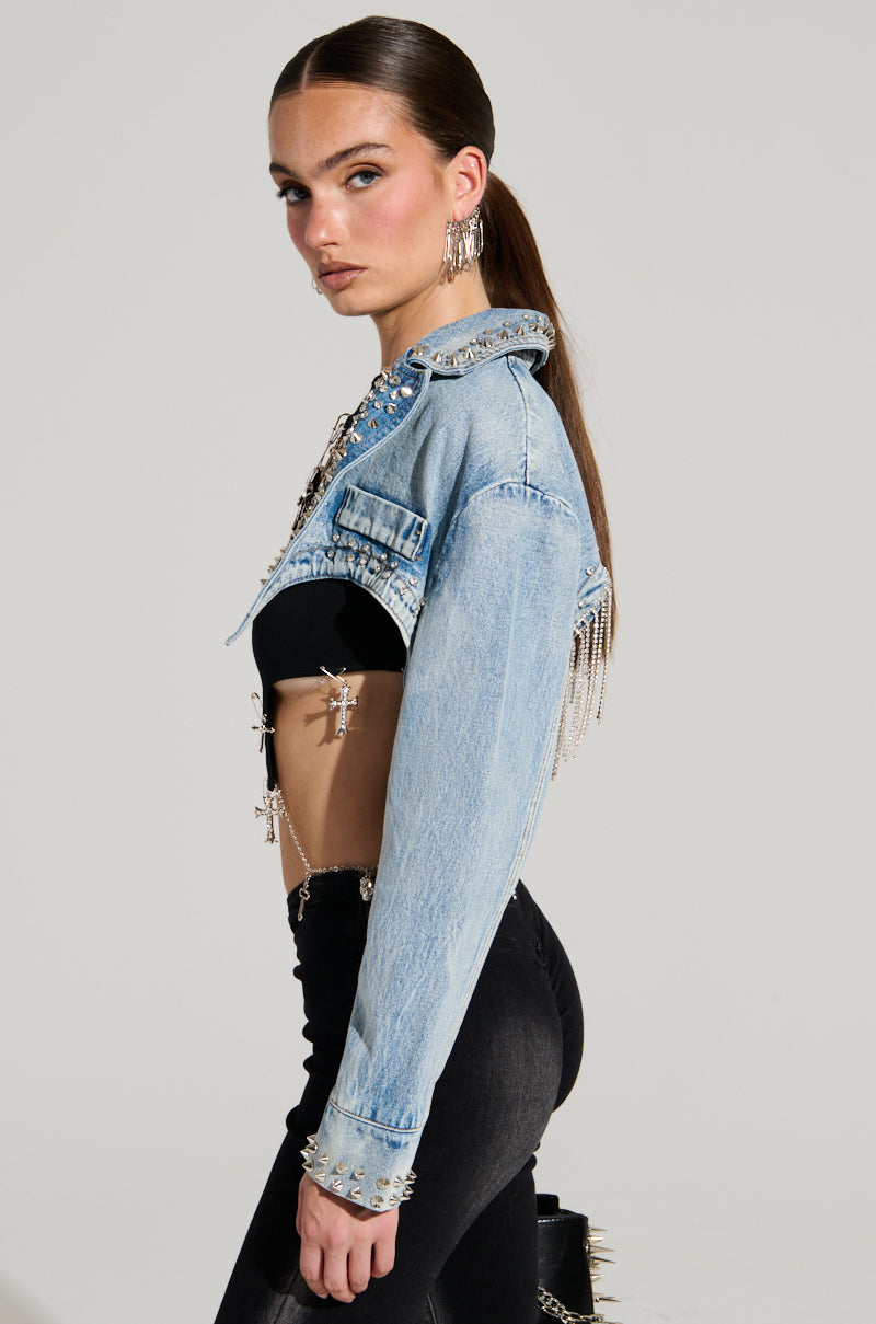 A person with long hair stands sideways, wearing the WATERFALL RHINESTONE CROPPED DENIM JACKET that features studs and fringe over a black top and high-waisted pants. They complete their look with large dangling earrings against a plain background.