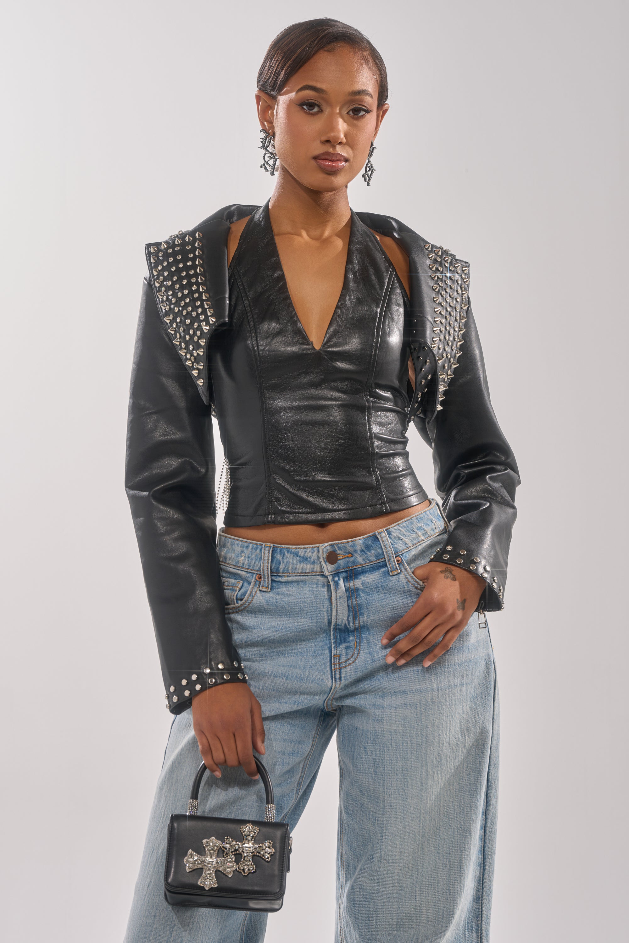 A woman with short hair and statement earrings models the NAOMI EMBELLISHED ULTRA CROP MOTO jacket, styled with structured shoulders, light blue wide-leg jeans, and a small black purse featuring silver accents. 0