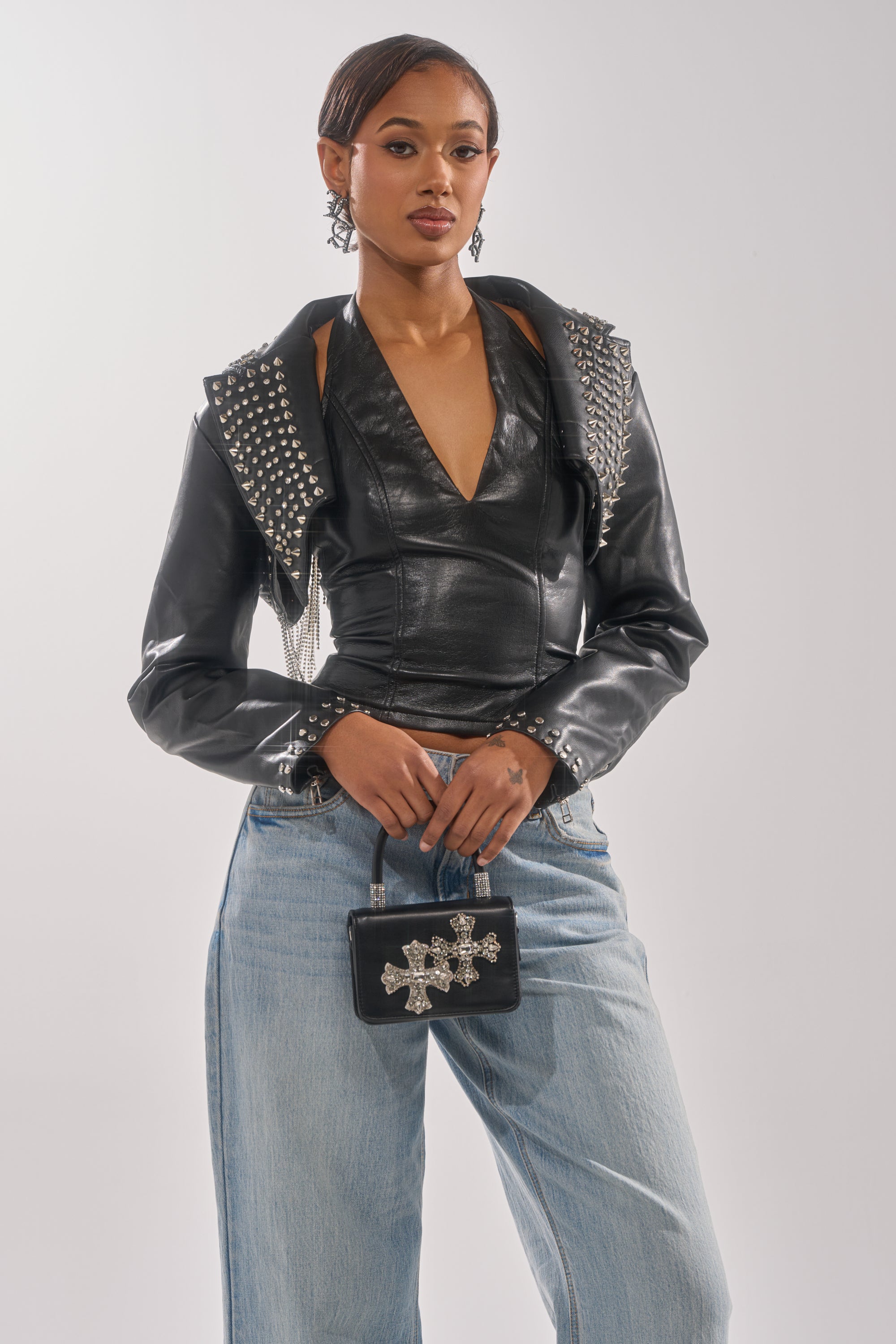 A woman poses confidently in the NAOMI EMBELLISHED ULTRA CROP MOTO jacket, paired with a black halter top, light blue jeans, and a small black handbag with silver embellishments against a plain light background. 1