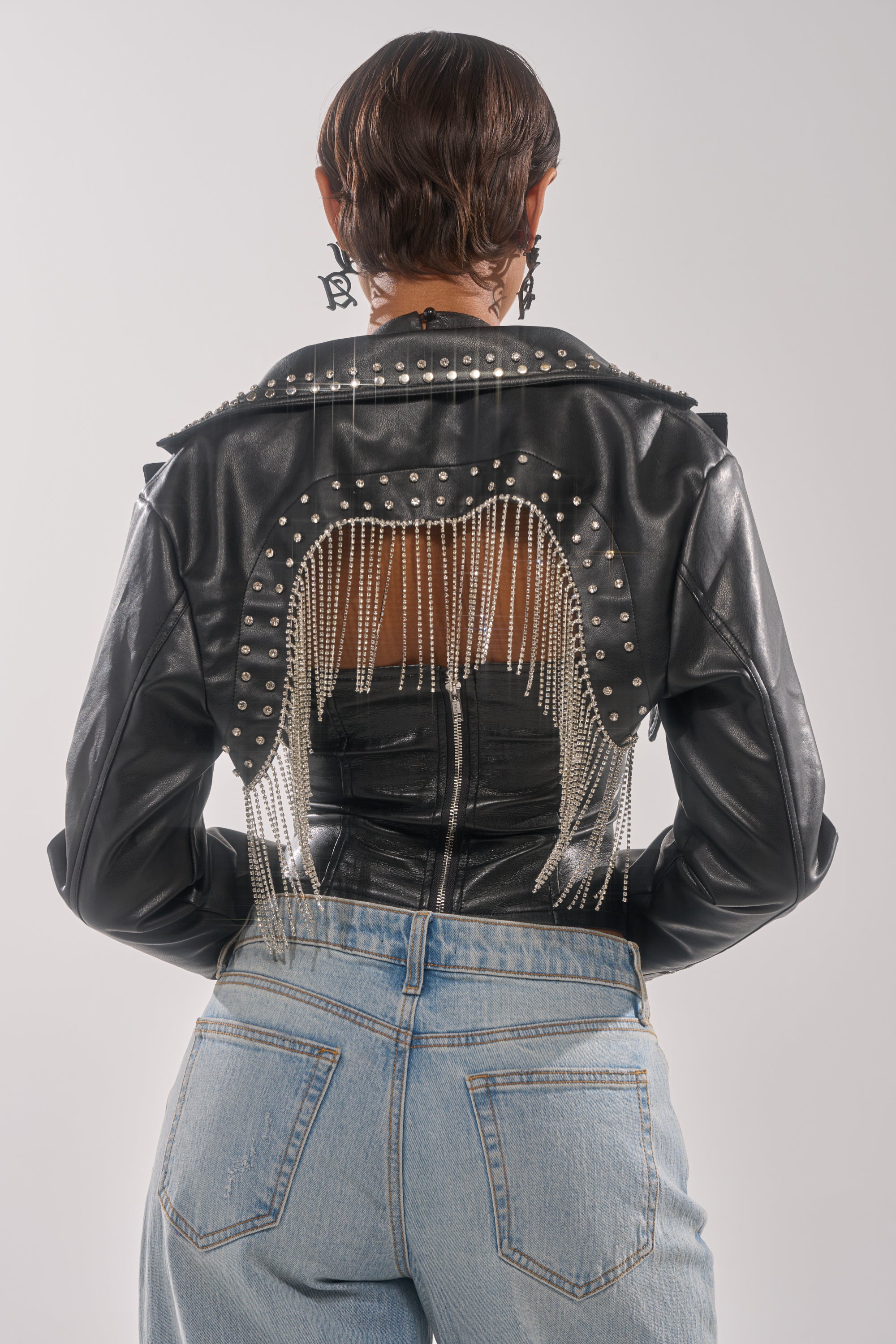 A person faces away, wearing the NAOMI EMBELLISHED ULTRA CROP MOTO jacket with silver studs, hanging chains, and a large back cutout, paired with light blue jeans. 3