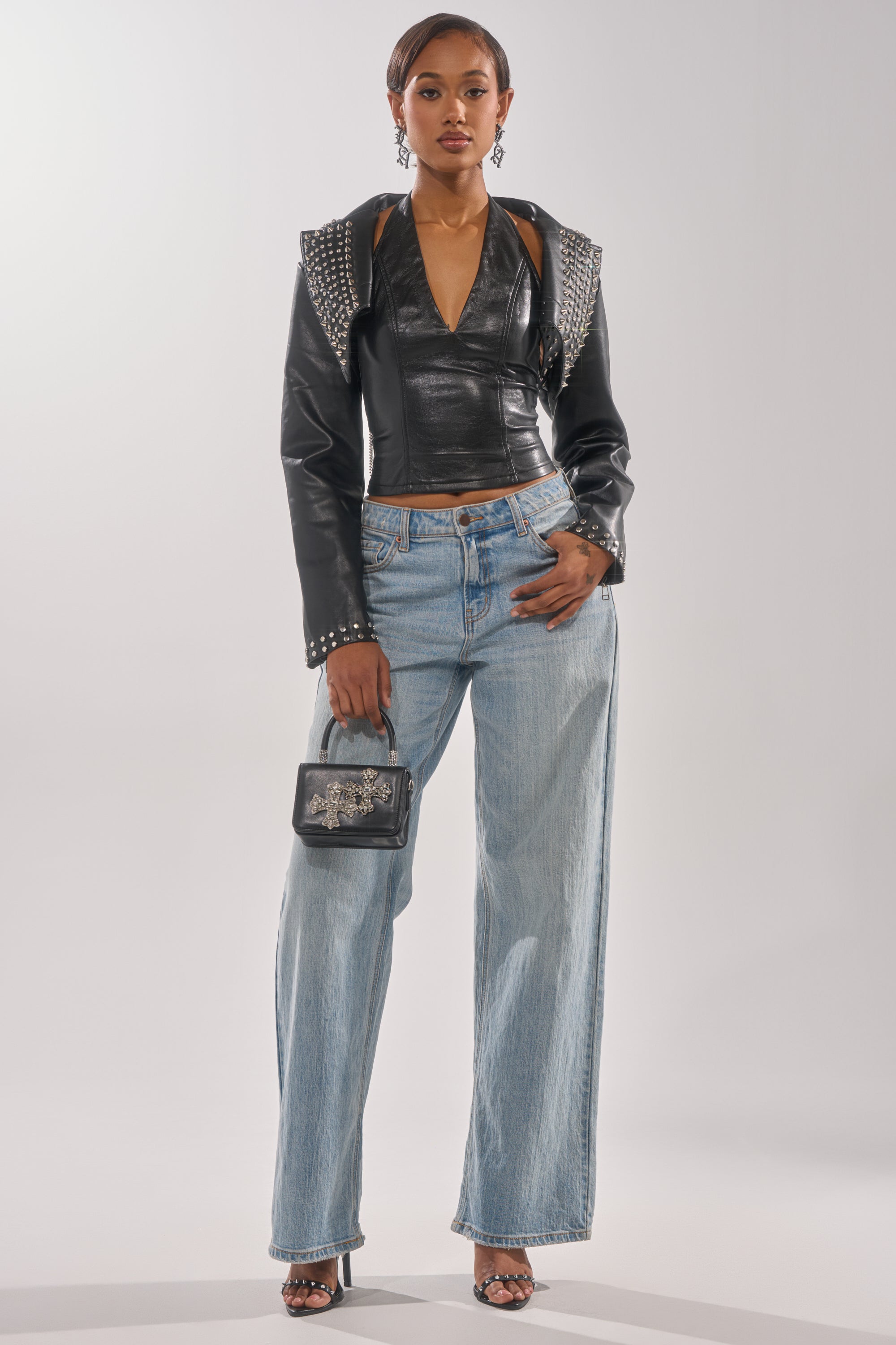 A woman stands confidently in the NAOMI EMBELLISHED ULTRA CROP MOTO jacket, paired with a black halter top, light blue wide-leg jeans, black heels, a small black handbag, and statement earrings. 4