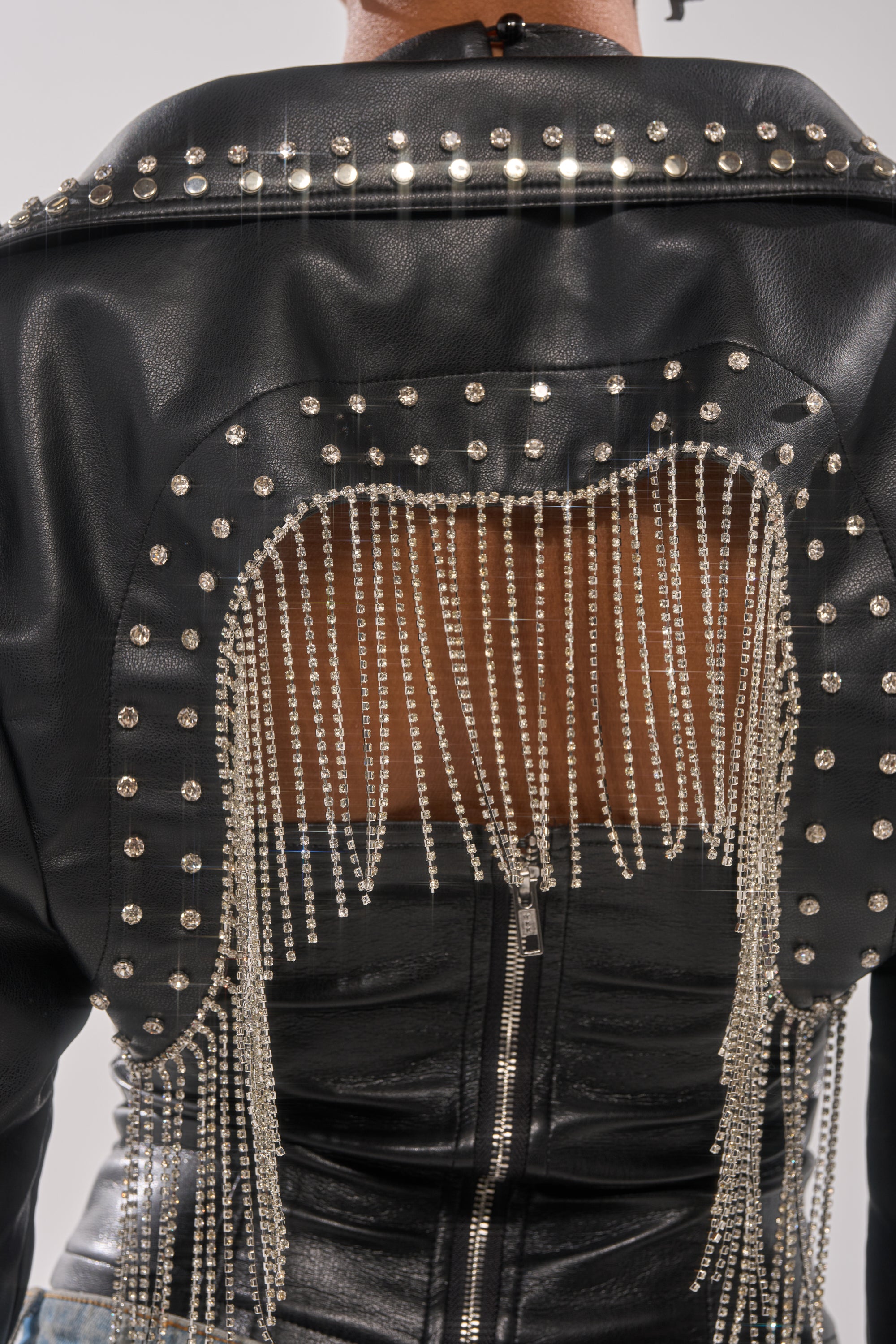A close-up of the back of the NAOMI EMBELLISHED ULTRA CROP MOTO, a faux leather jacket with a cutout design accented by metallic studs and long, silver chain fringe. 5