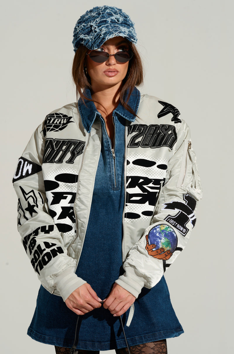 Modeling the UNITY BOMBER, an individual exudes confidence in front of a neutral backdrop. Their ensemble includes a denim dress, sleek sunglasses, and a textured blue hat, perfectly complementing the white and black graphic bomber jacket embellished with embroidered patches. 0