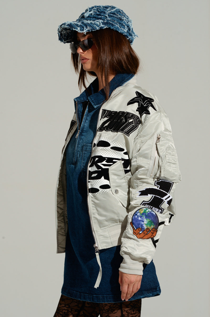 A woman wearing the UNITY BOMBER, a white jacket embellished with embroidered patches, paired with a denim dress, sunglasses, and a blue denim hat. She has long hair and is facing sideways against a neutral background. 2