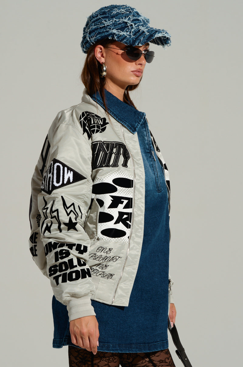 A person wearing the UNITY BOMBER graffiti-print windbreaker, a denim dress, a furry blue cap, sunglasses, and hoop earrings stands against a plain background. 3