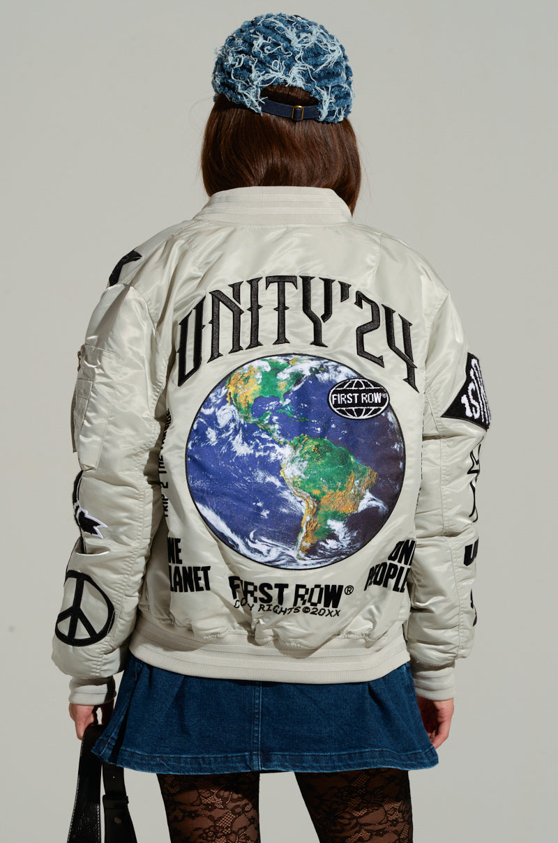 Wearing the UNITY BOMBER jacket, embellished with embroidered patches featuring a large globe and slogans such as "UNITY '24" and "ONE PLANET ONE POPULATION," a person with long hair completes their look with a blue patterned beanie, denim skirt, and lace-patterned leggings. 4