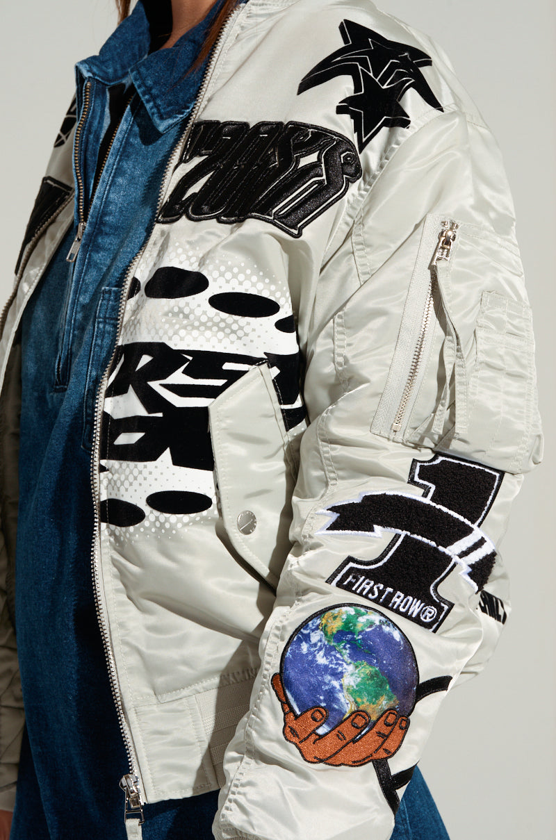 A person showcasing the UNITY BOMBER, a white jacket featuring an array of black embroidered patches and logos, including an earth patch on the sleeve, pairs it stylishly with a blue denim shirt underneath. 7