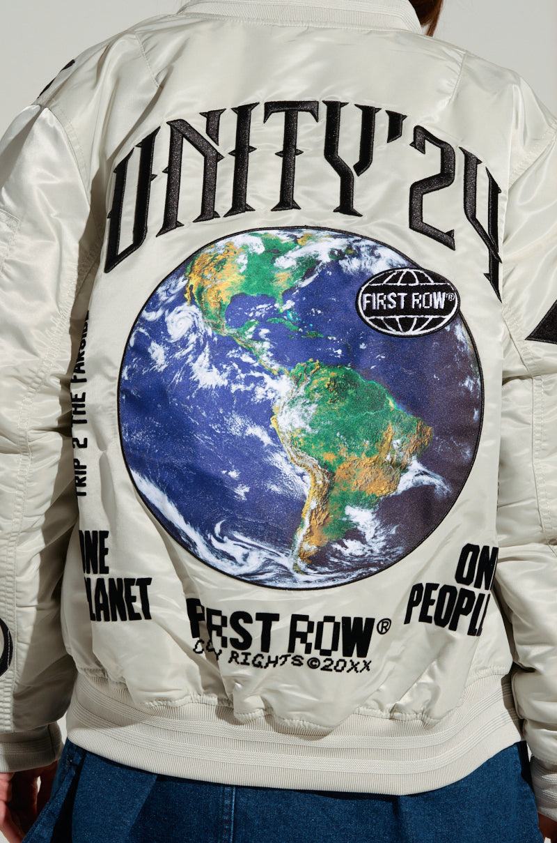 An individual wearing the UNITY BOMBER, a light-colored jacket highlighted by a prominent image of Earth. The jacket is embellished with embroidered patches and features text such as "UNITY '24," "FIRST ROW," "ONE PLANET," and "ONE PEOPLE." The person stands with part of their jeans visible. 8