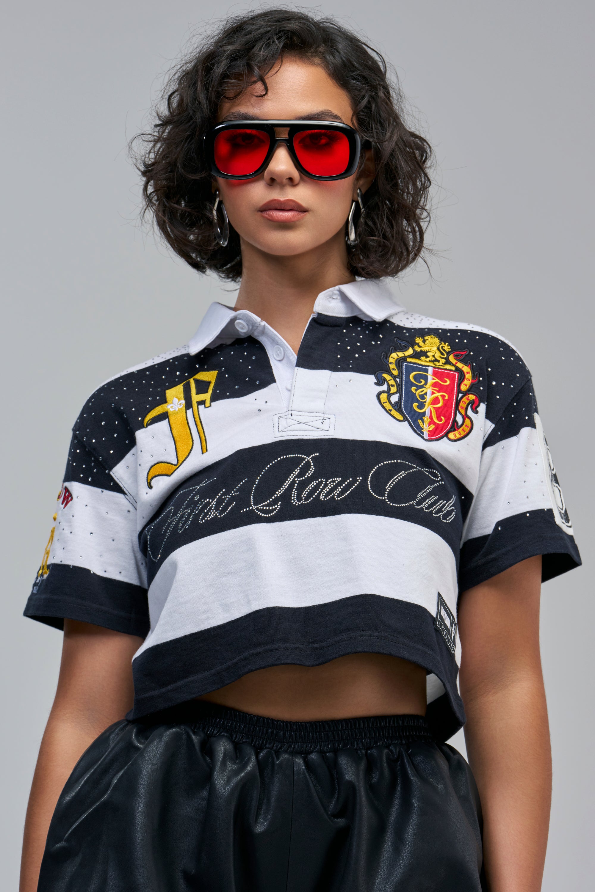 A person wearing a FIRST ROW CLUB STRIPED TEE, a cropped black and white striped polo with a cursive logo, an embroidered crest, a large varsity-style letter J on the chest, and black sunglasses.