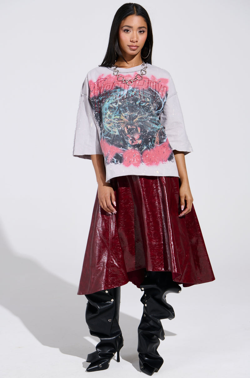 Wearing the PRIMAL FURY CROPPED GRAPHIC SWEATSHIRT with an abstract design, paired with a shiny dark red asymmetrical skirt and tall black boots, they pose confidently against a plain backdrop, exuding vintage-inspired jaguar grace.