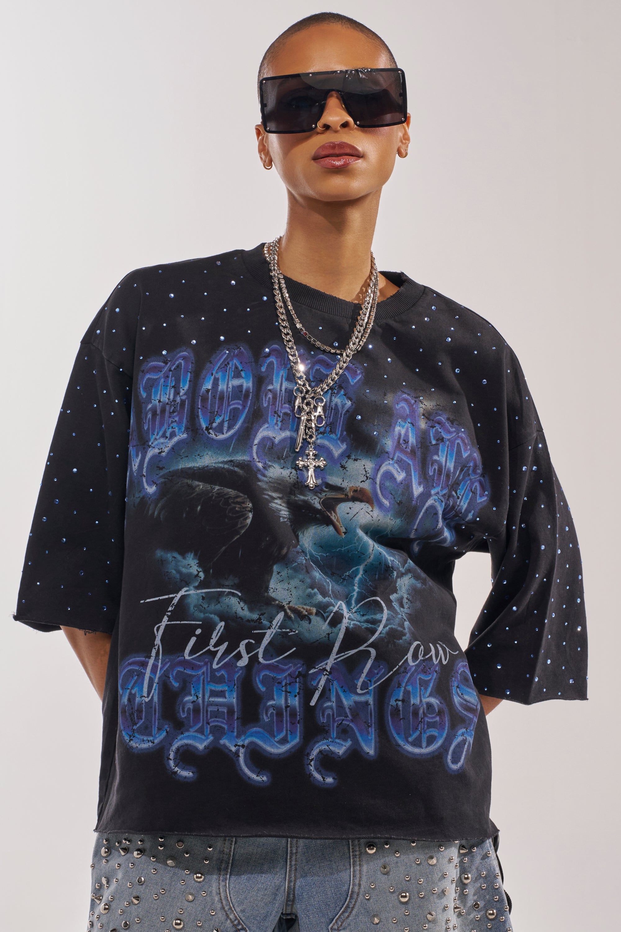A person with a shaved head and large black sunglasses wears the FIRST ROW TAKING FLIGHT FRENCH TERRY TEE with blue gothic lettering, layered silver necklaces, and studded denim shorts, standing against a plain background. 0