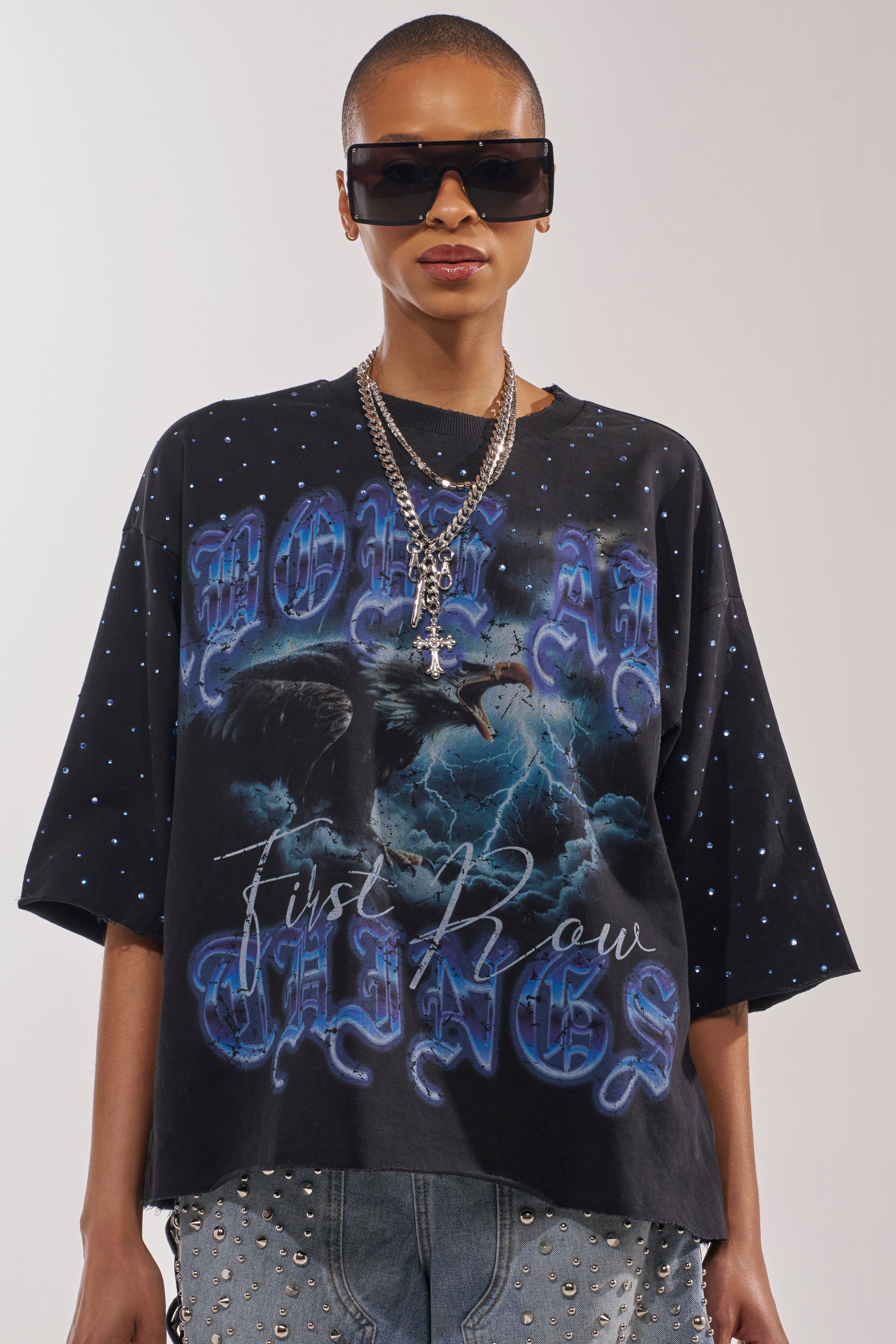 A person with a shaved head wears oversized black sunglasses, layered chain necklaces, and the FIRST ROW TAKING FLIGHT FRENCH TERRY TEE— a gothic black graphic shirt featuring blue gothic text, raven graphics, and white dots. 1
