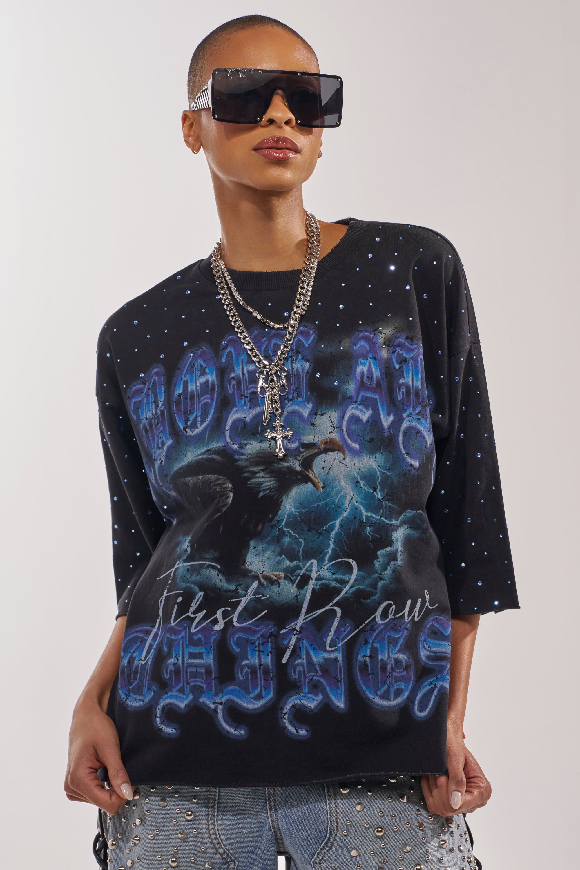 A person with short hair and a confident pose wears large black sunglasses, layered silver necklaces, and the FIRST ROW TAKING FLIGHT FRENCH TERRY TEE—a gothic black graphic tee with blue designs. 2