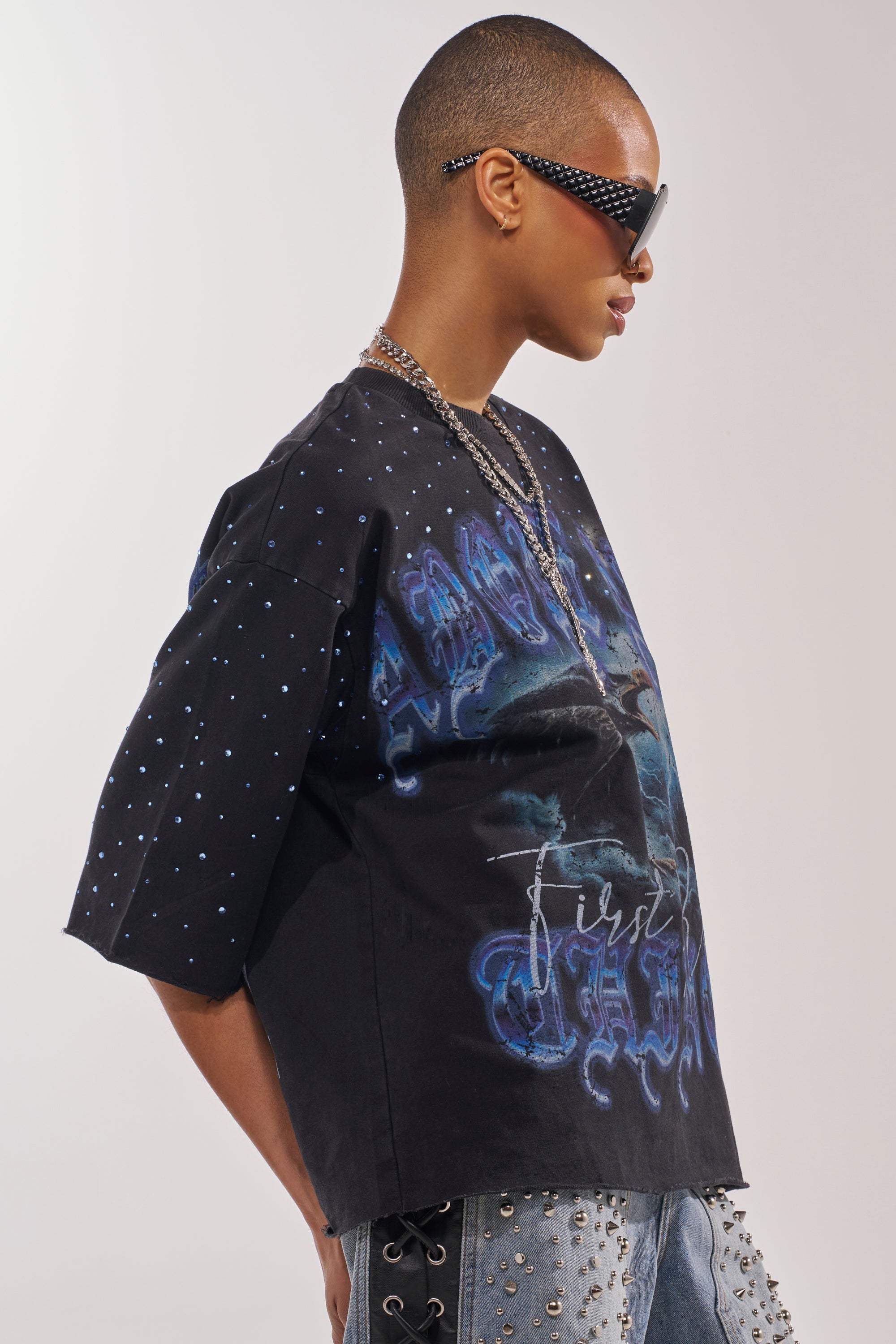 A person with a shaved head and studded sunglasses poses in profile against a plain background, wearing the FIRST ROW TAKING FLIGHT FRENCH TERRY TEE, silver chains, and studded pants. 3