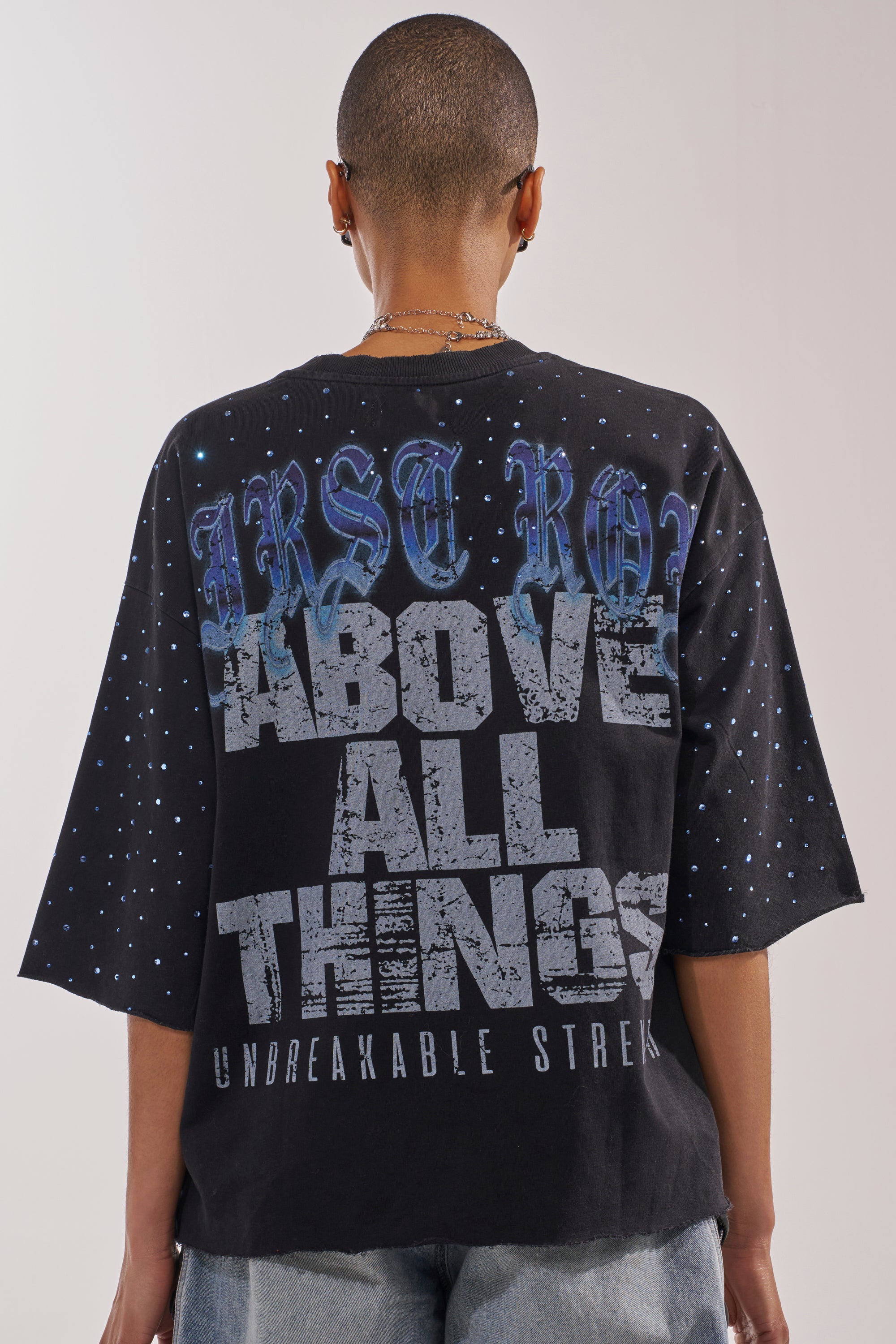 Seen from behind, a person with short hair wears the FIRST ROW TAKING FLIGHT FRENCH TERRY TEE—a black graphic shirt featuring blue and white speckles and large "FIRST ROW ABOVE ALL THINGS UNBREAKABLE STREAK" text for bold streetwear appeal. 4