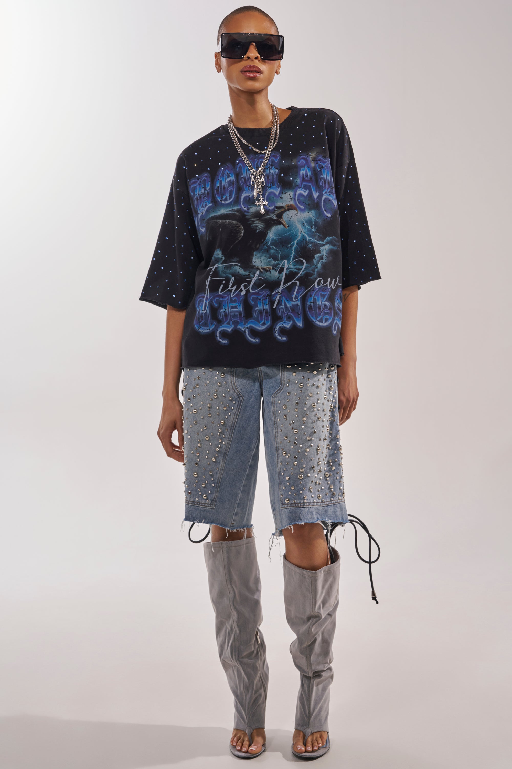 A person in oversized sunglasses, studded denim shorts, layered necklaces, tall gray open-toe boots, and the FIRST ROW TAKING FLIGHT FRENCH TERRY TEE with blue designs stands against a plain background. 5