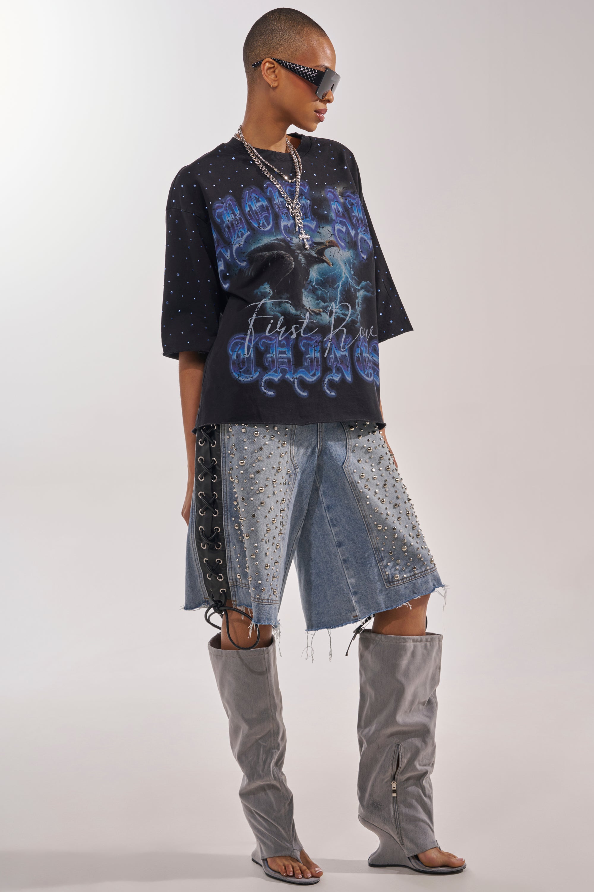 A person in sunglasses, the FIRST ROW TAKING FLIGHT FRENCH TERRY TEE, studded denim shorts with side lacing, layered necklaces, and tall gray slouchy open-toe boots stands against a plain light background. 6