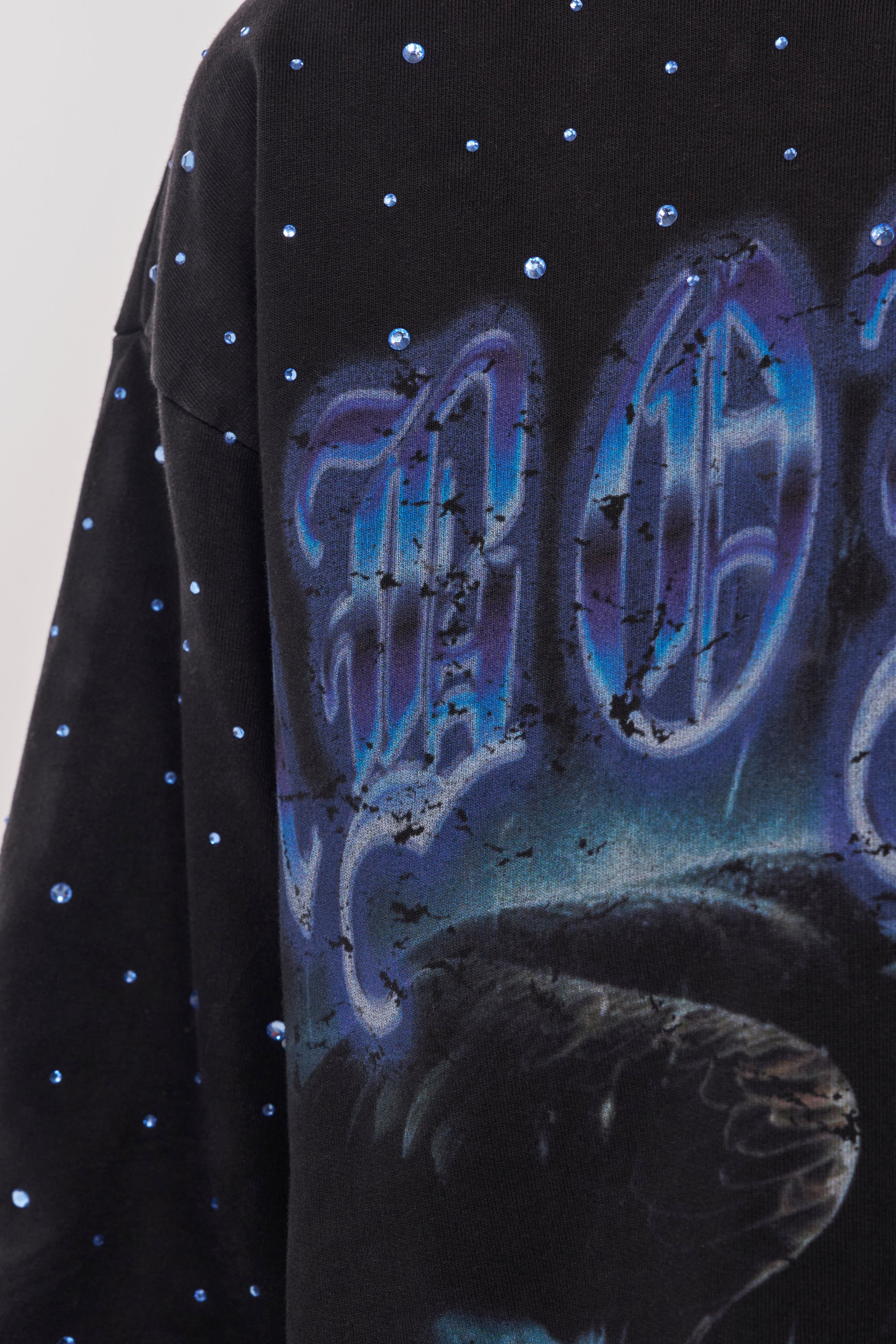 A close-up of the FIRST ROW TAKING FLIGHT FRENCH TERRY TEE, a black graphic shirt with blue rhinestone accents and a distressed blue design—ideal for adding edgy style to your streetwear wardrobe. 7