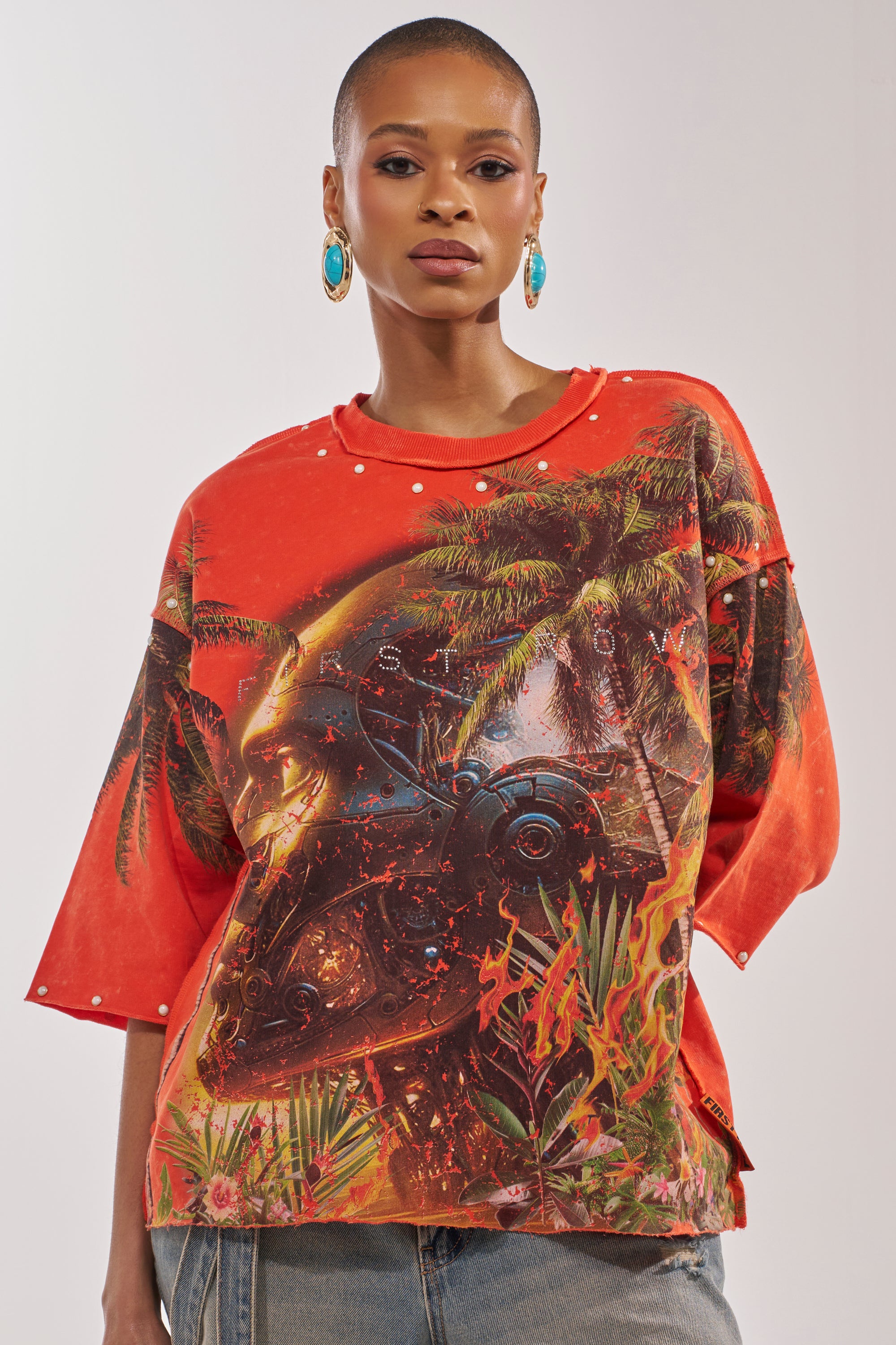 Wearing the FIRST ROW LOST PARADISE RHINESTONE FRENCH TERRY TEE with tropical palm tree and flame graphics, a short-haired person in blue earrings pairs it with light denim jeans against a plain background. 0