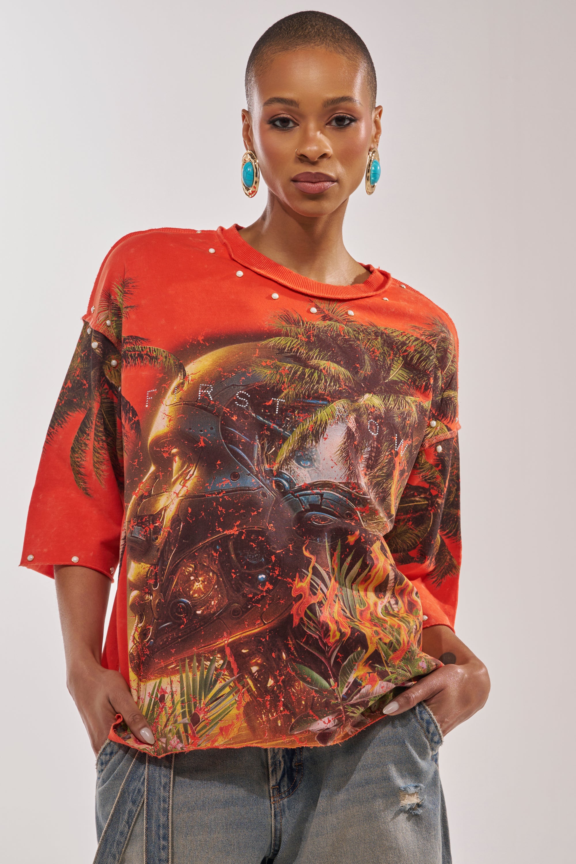 A woman with short hair and turquoise earrings wears the FIRST ROW LOST PARADISE RHINESTONE FRENCH TERRY TEE featuring a surreal graphic and pearl studs, paired with baggy denim jeans, standing confidently with hands in her pockets. 1