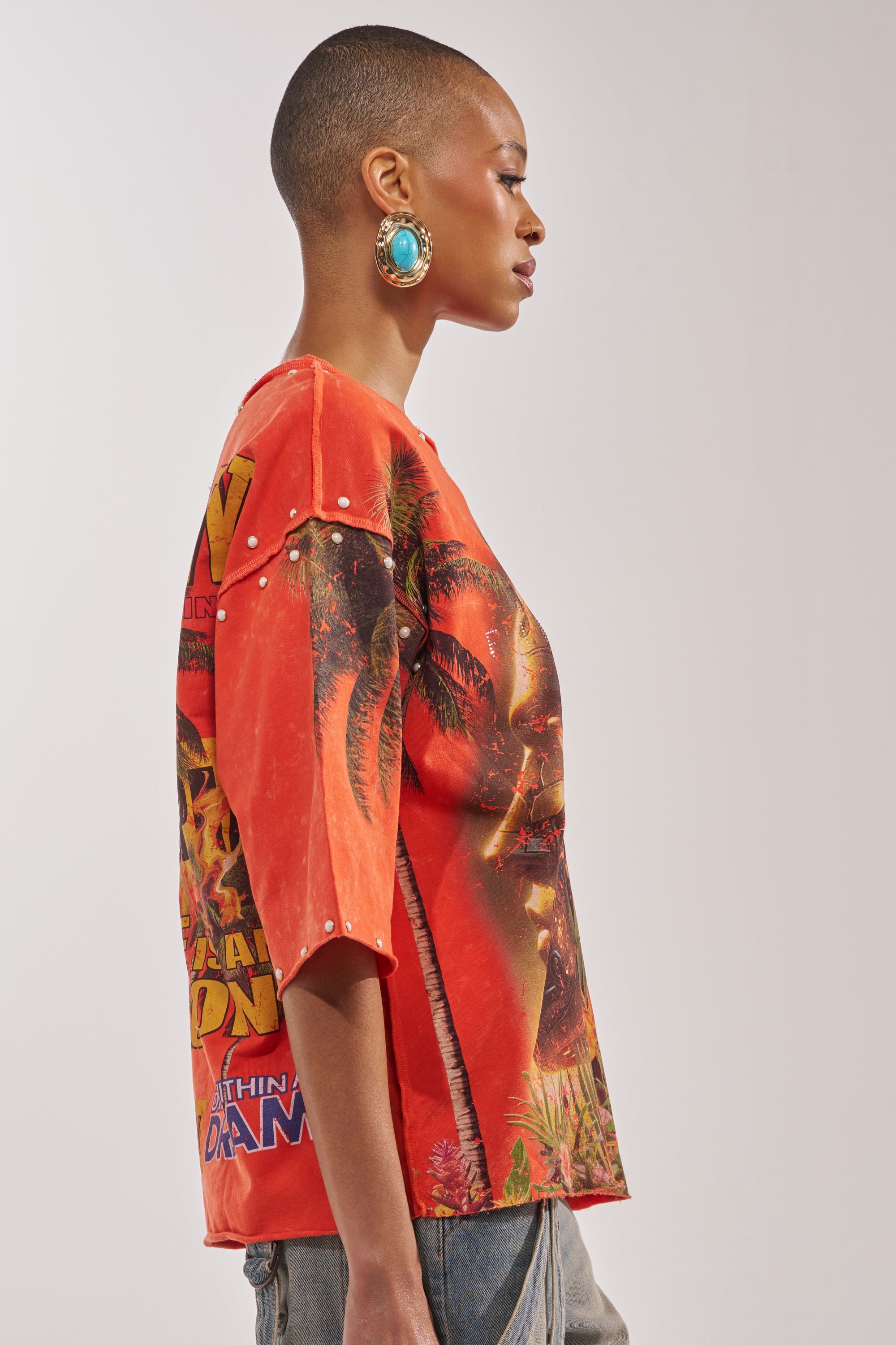 A person with a shaved head and large turquoise earrings wears the FIRST ROW LOST PARADISE RHINESTONE FRENCH TERRY TEE in orange, styled with blue jeans, standing in profile against a plain background. 2