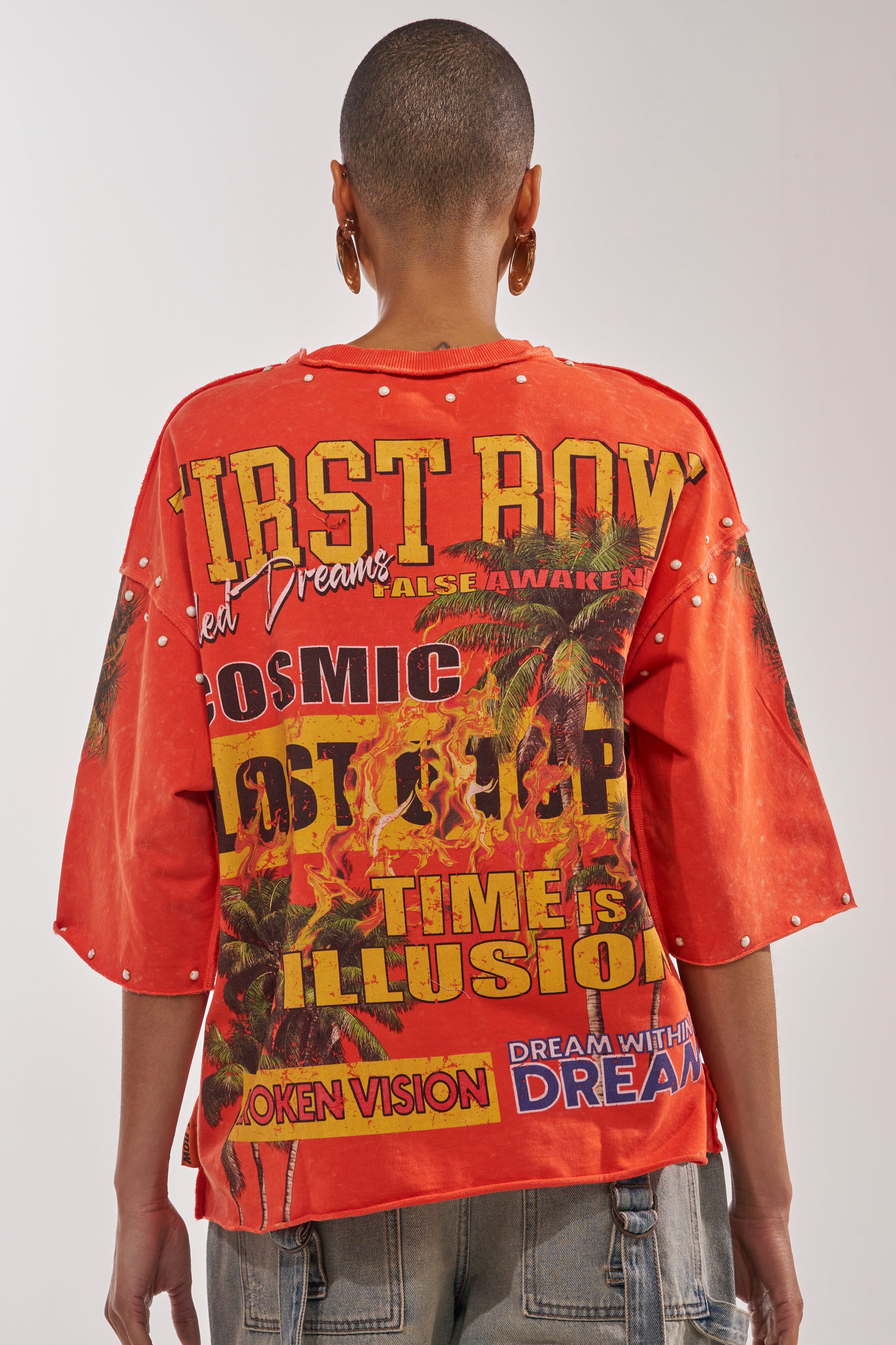 A person with a shaved head wears the FIRST ROW LOST PARADISE RHINESTONE FRENCH TERRY TEE—an eye-catching orange shirt featuring bold, colorful palm tree graphics and surreal text like "FIRST ROW," "COSMIC," and "TIME IS ILLUSION. 3