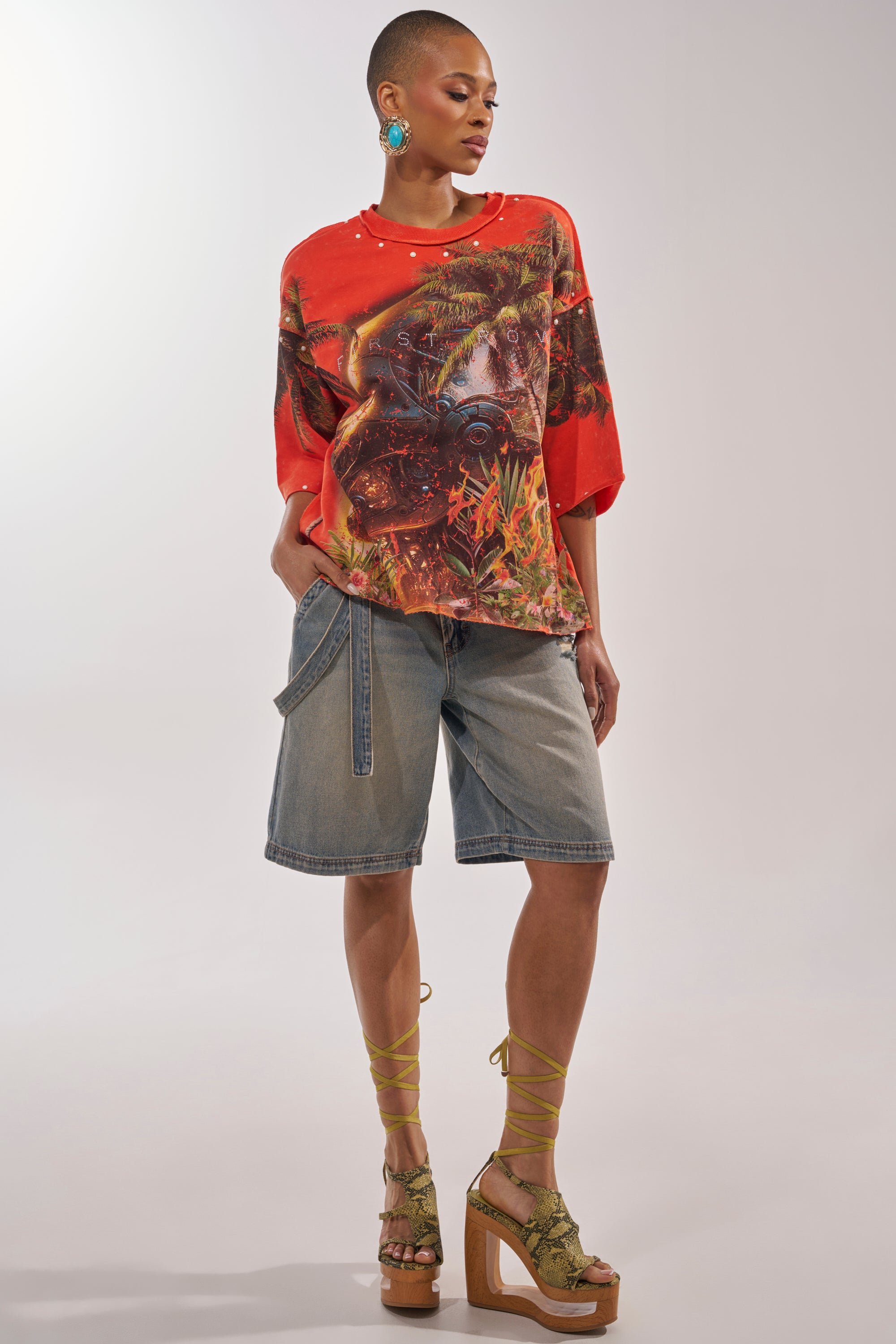 A person wears the FIRST ROW LOST PARADISE RHINESTONE FRENCH TERRY TEE, loose denim Bermuda shorts, green lace-up platform sandals, and turquoise statement earrings while standing confidently against a plain background. 4